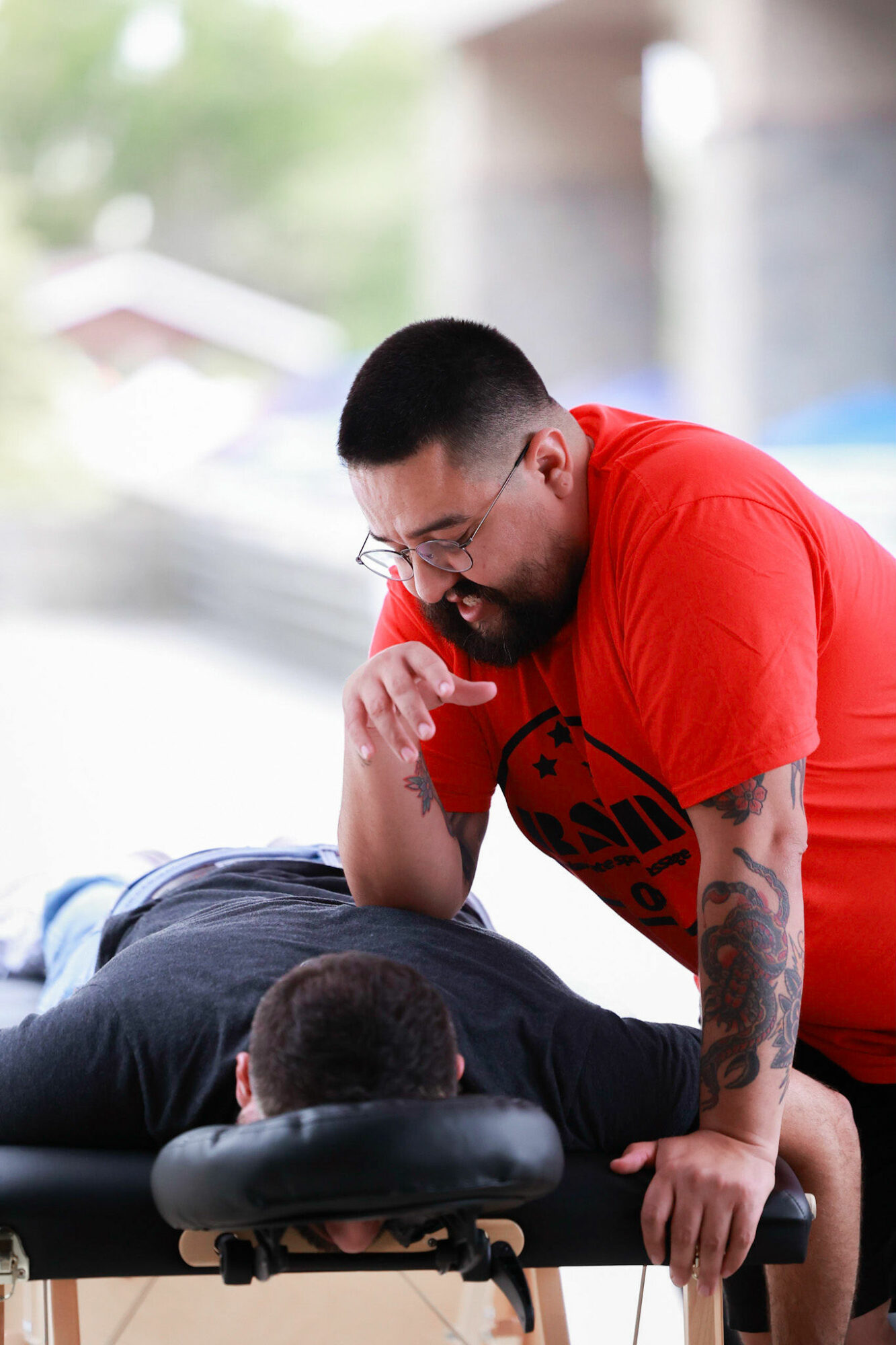 Man with glasses and tattoos performs a massage on a person lying face down outdoors, under a bridge or structure.