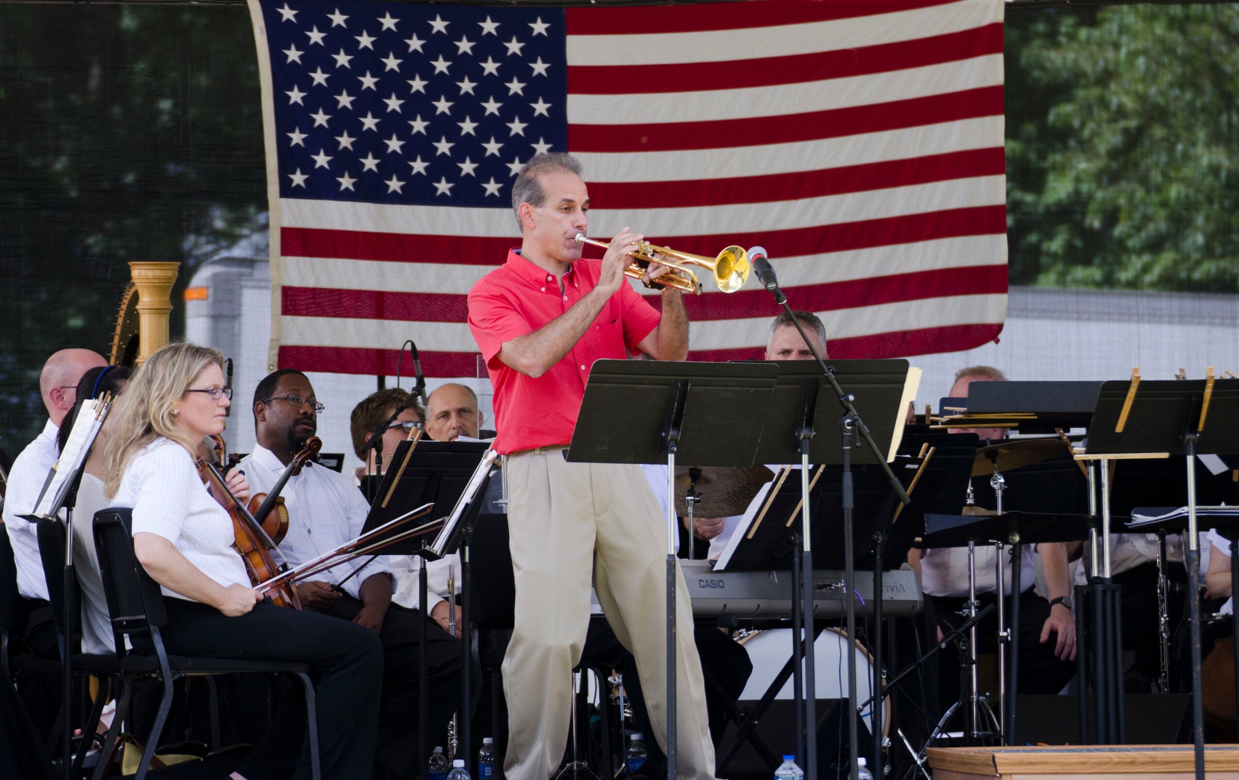 A young man plays the trumpet in a band with children, with an American flag in the background outdoors.