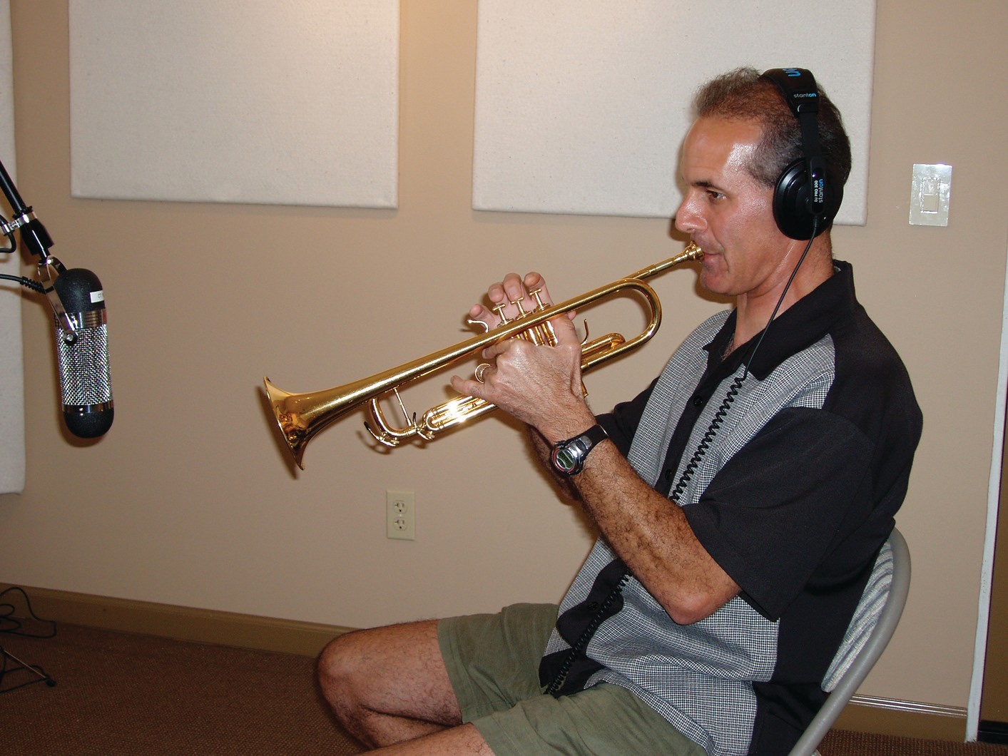 Man playing trumpet in recording studio, wearing headphones, sitting on chair, microphone nearby, beige wall background.