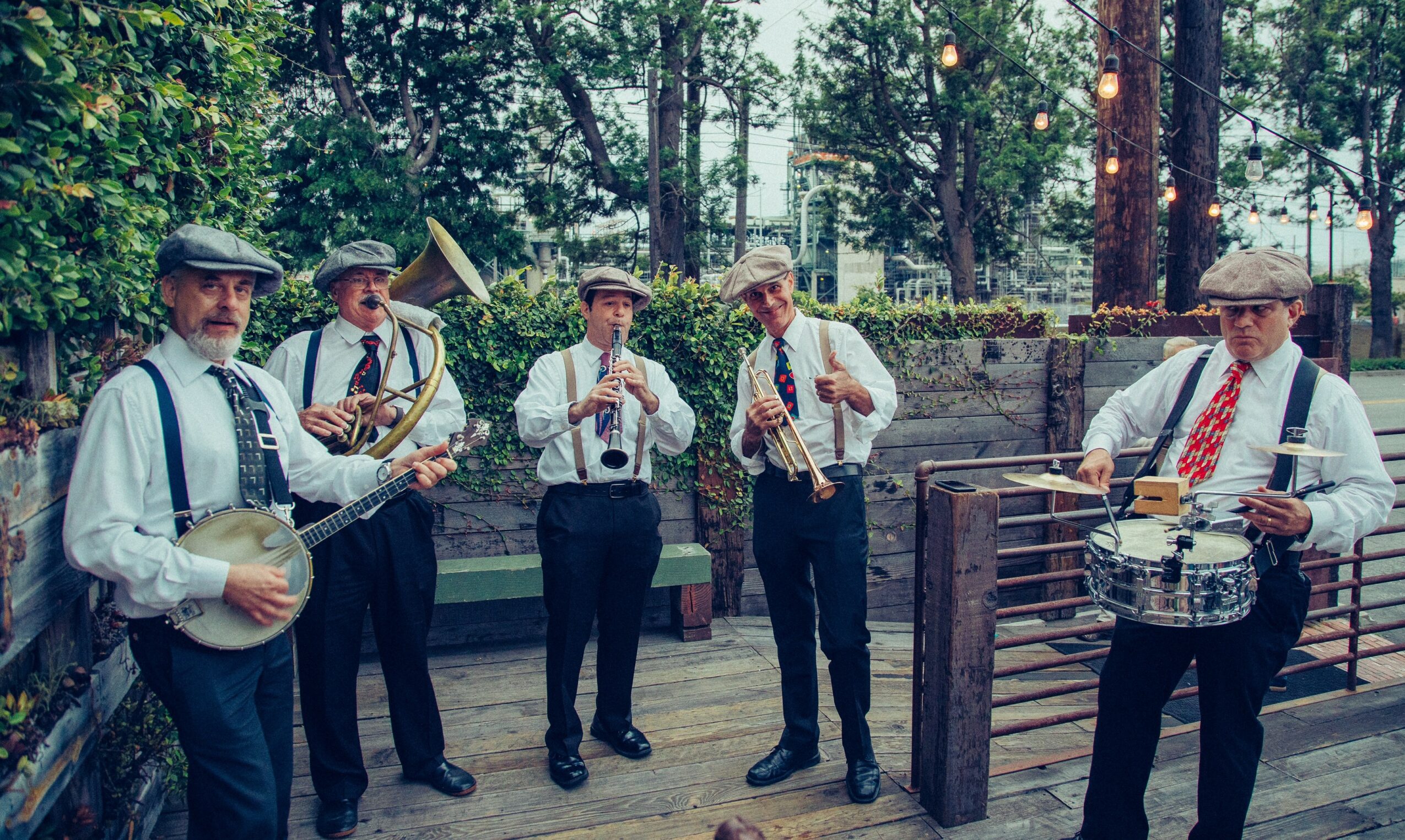 Six people playing musical instruments outdoors, surrounded by trees and wooden fencing.