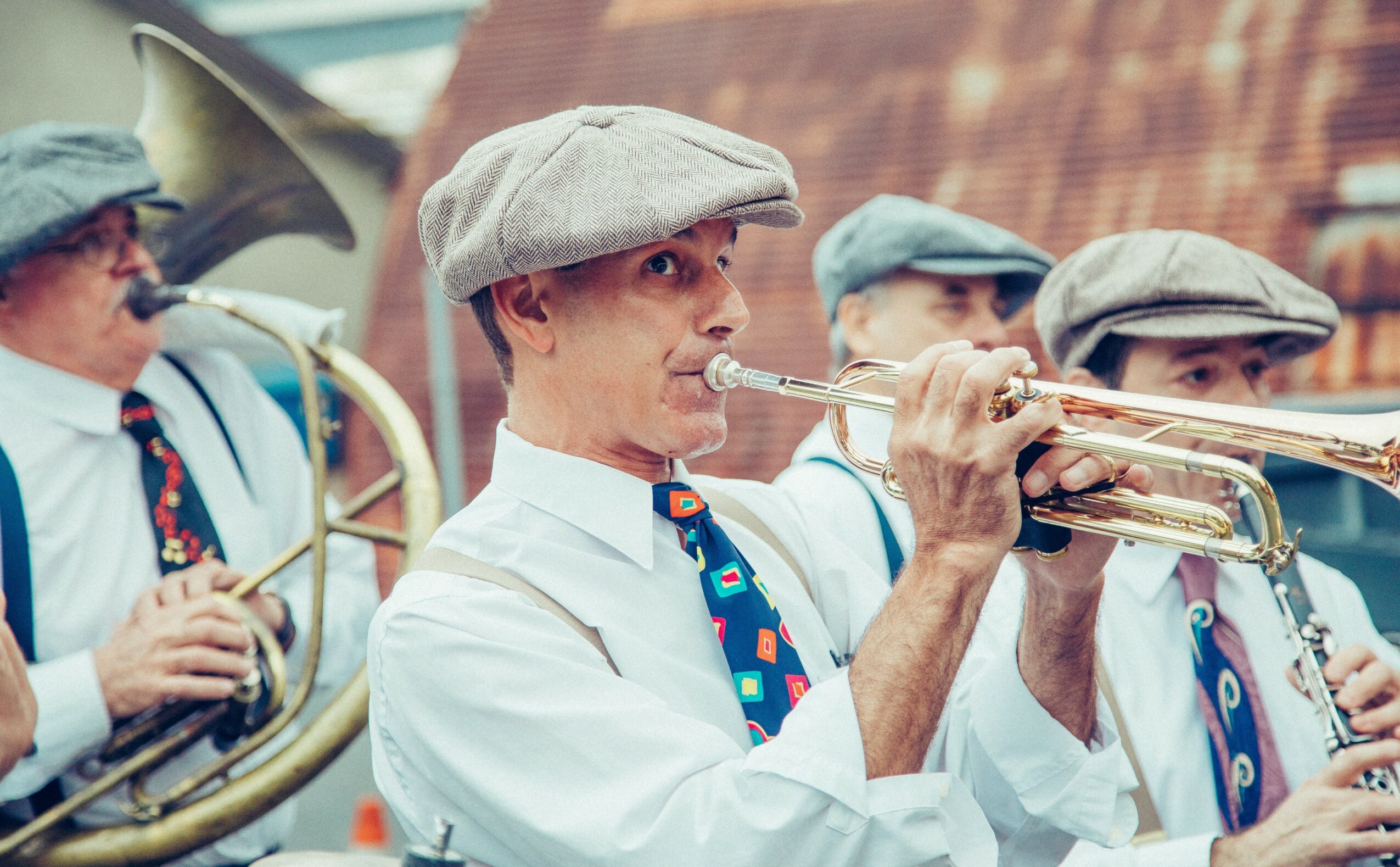 Group of musicians playing brass instruments outdoors, wearing hats and white shirts, with a brick building in the background.