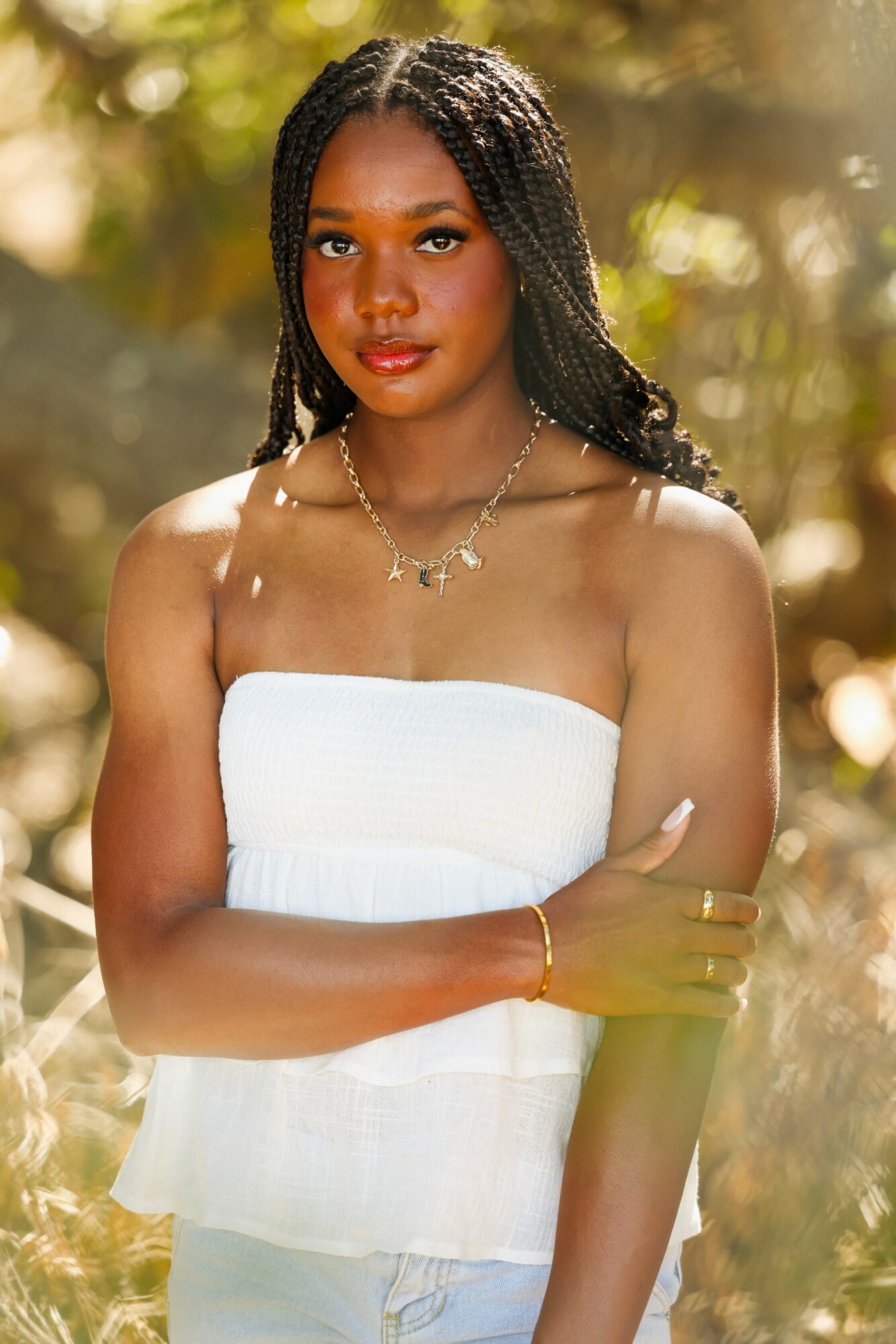 Young woman with braided hair wearing a white strapless dress outdoors, sunlight filtering through trees, arms crossed, jewelry visible.