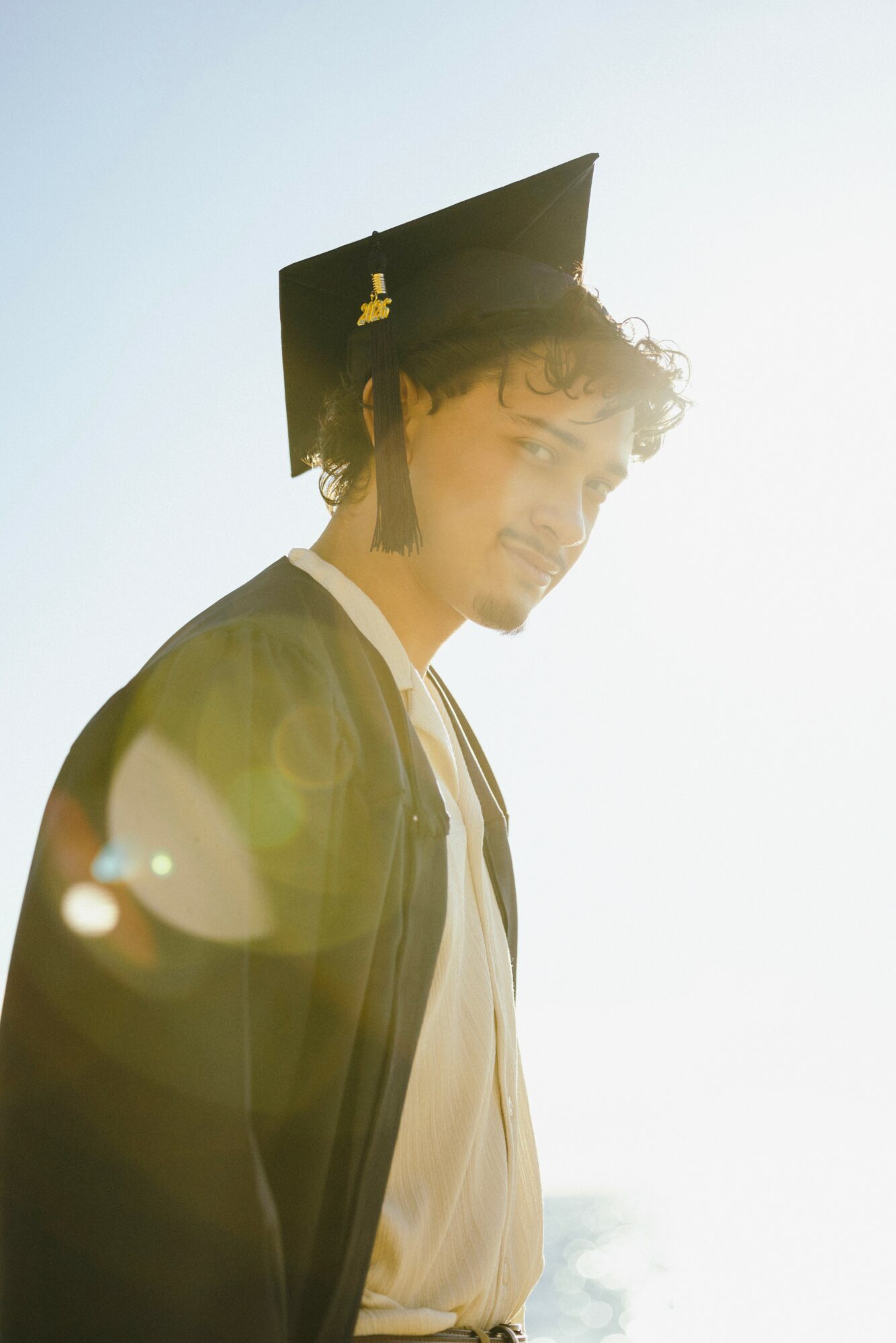 Young man in graduation cap and gown smiling, sunlight behind him, lens flare visible.