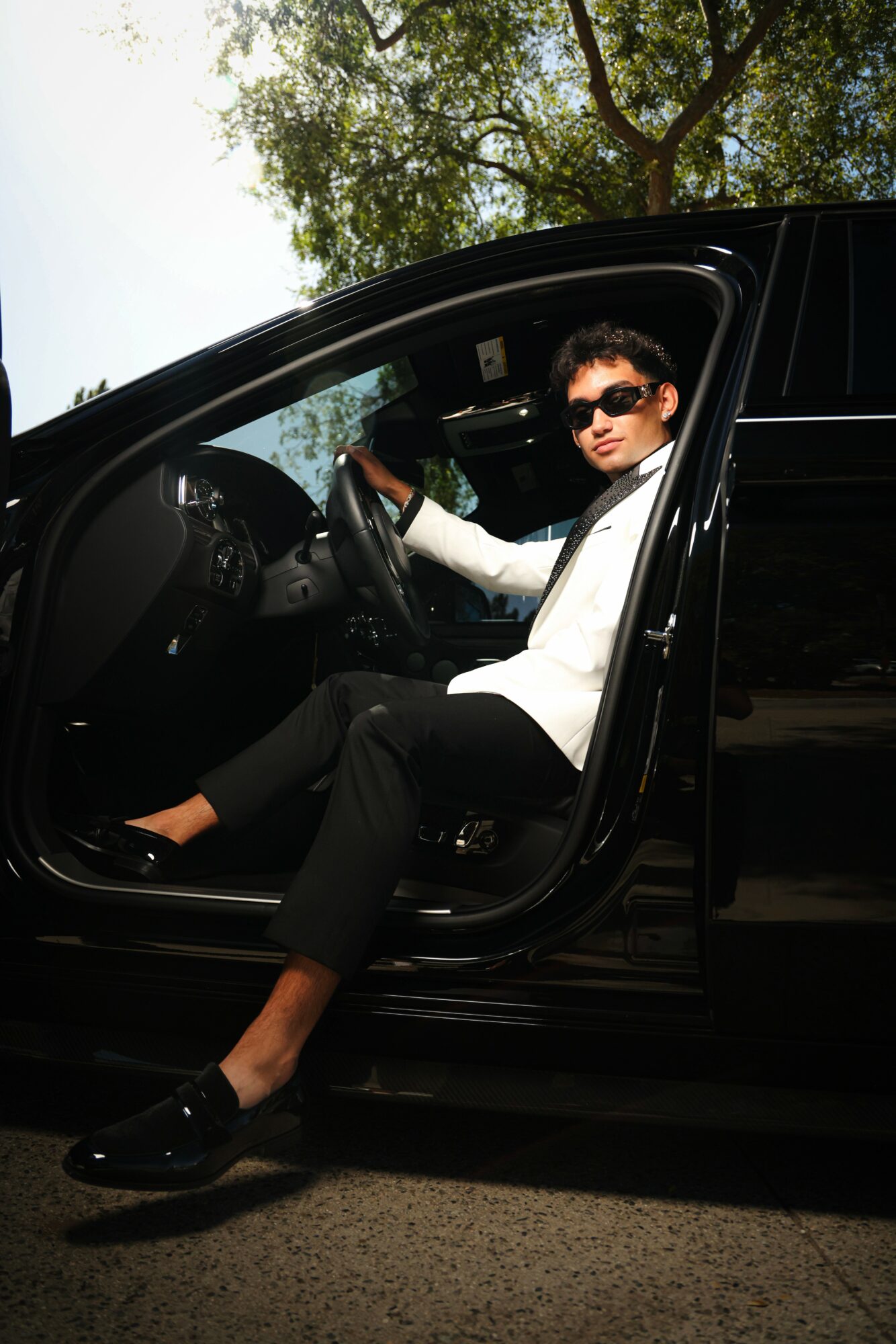 Woman sitting in car with open door, wearing sunglasses, white blazer, black pants, and black shoes, outdoors with trees in background.