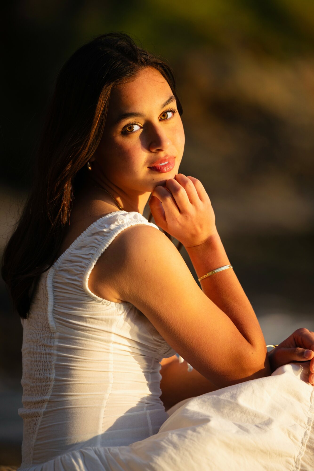 Young woman with long dark hair wearing a white sleeveless dress, sitting outdoors with a blurred background, looking at the camera.