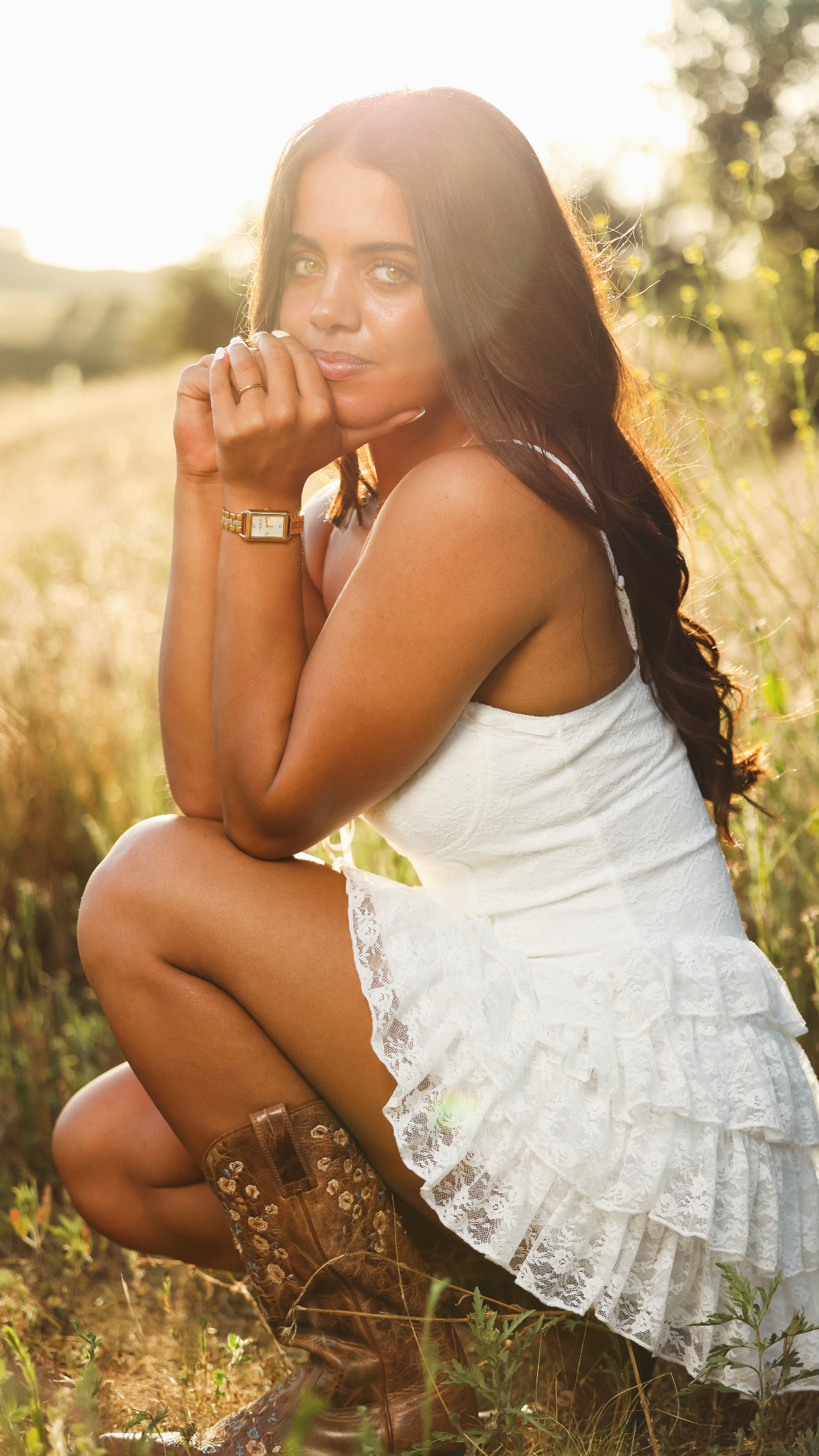 Young woman crouching outdoors in sunlight, wearing a white dress and brown boots, with long dark hair and a watch.