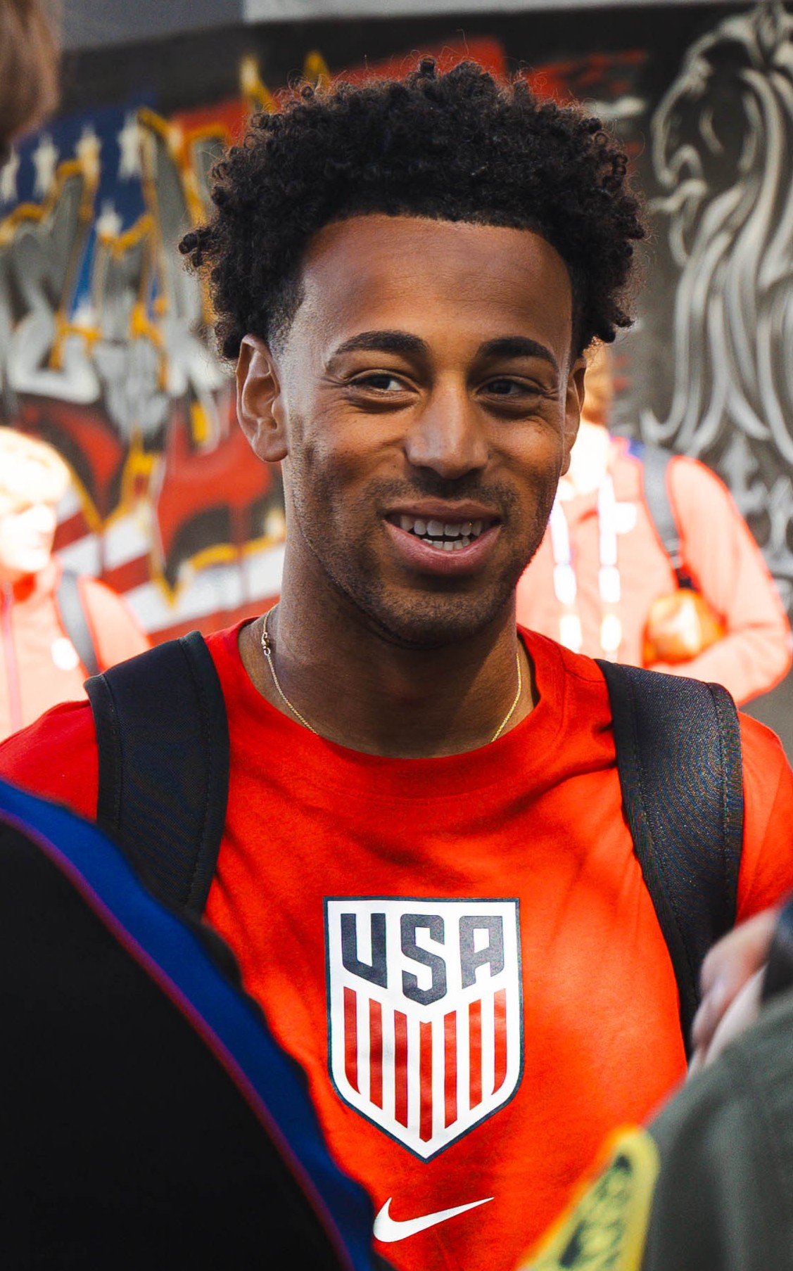 Young man with curly hair wearing a red USA shirt and backpack, smiling outdoors with graffiti background.