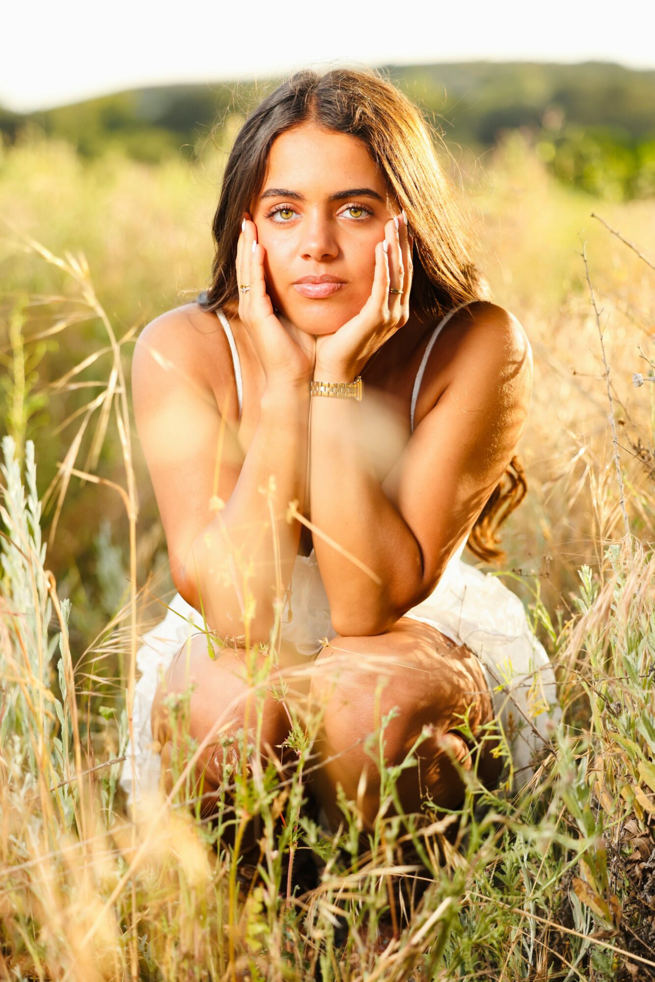 Young woman with long hair sitting in a grassy field, resting her chin on her hands, looking at the camera.
