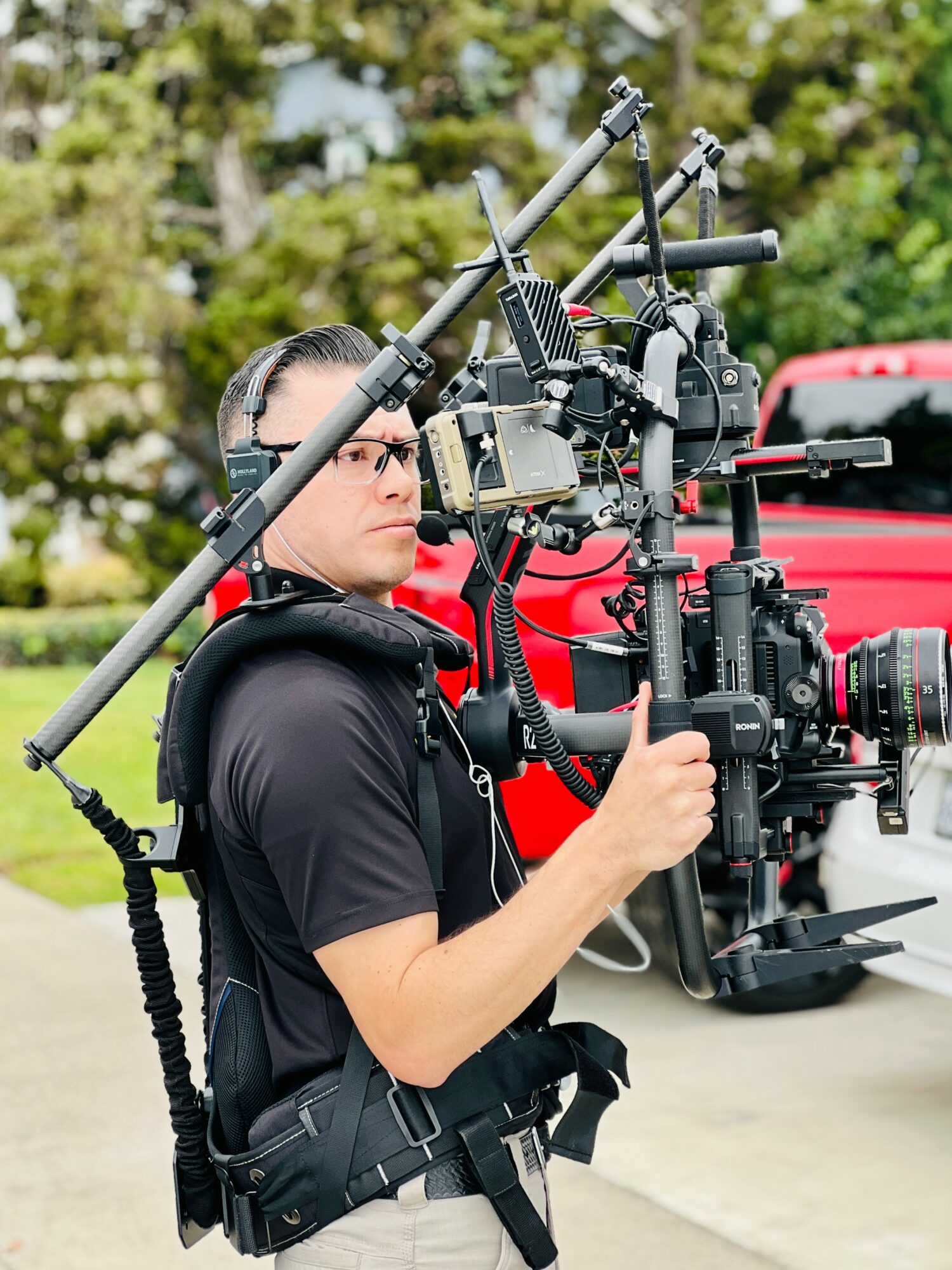 Person operating a professional camera mounted on a stabilizer outdoors, with trees and a red vehicle in the background.
