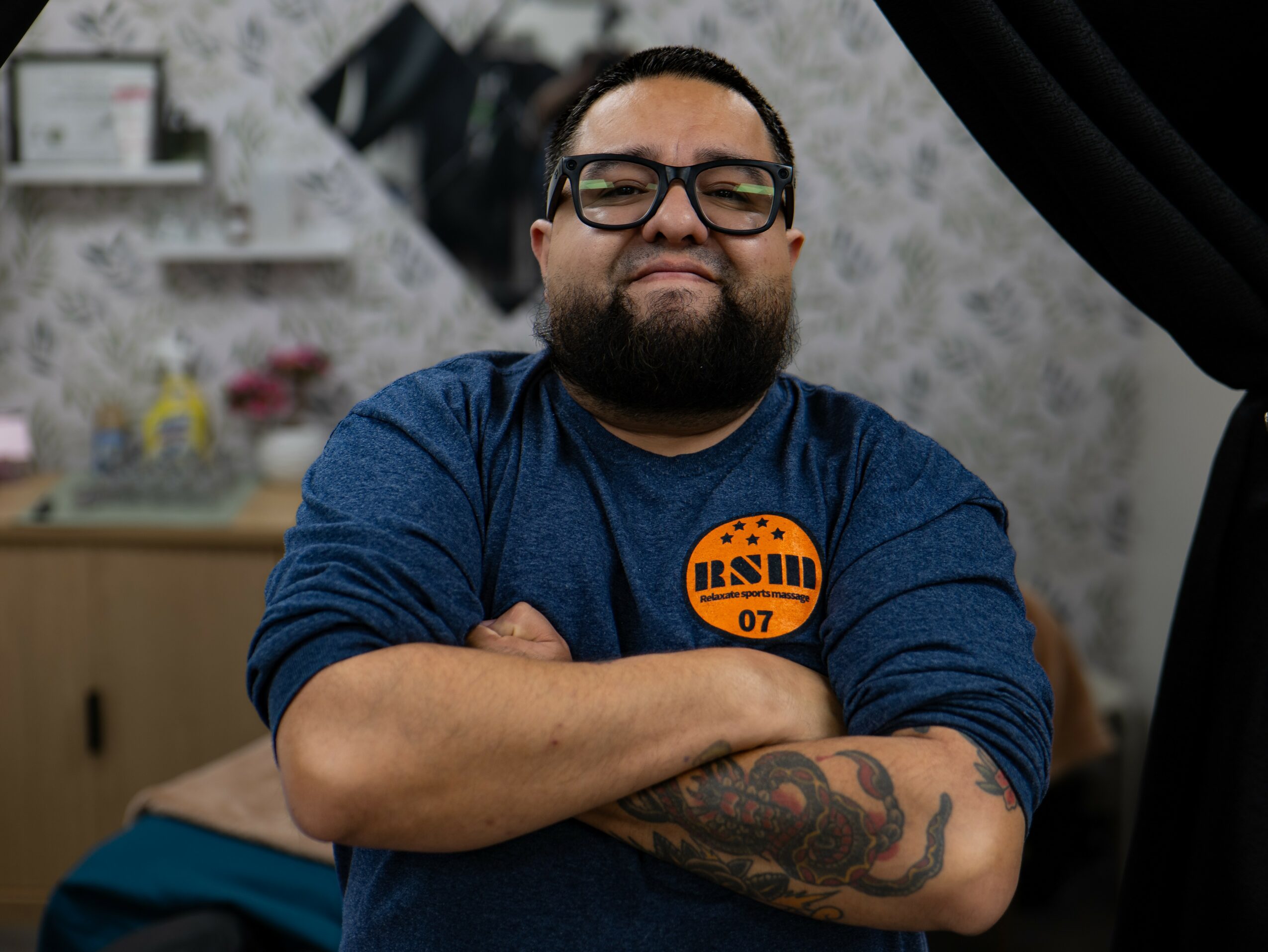 Man with glasses and a beard, arms crossed, standing indoors with a tattooed arm, smiling slightly.