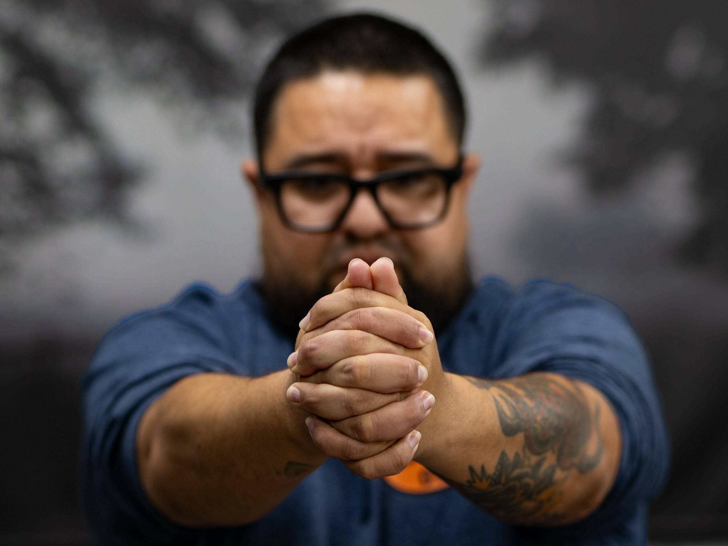 Man with glasses and a tattoo on his arm, clasping his hands together, outdoors with blurred trees in background.