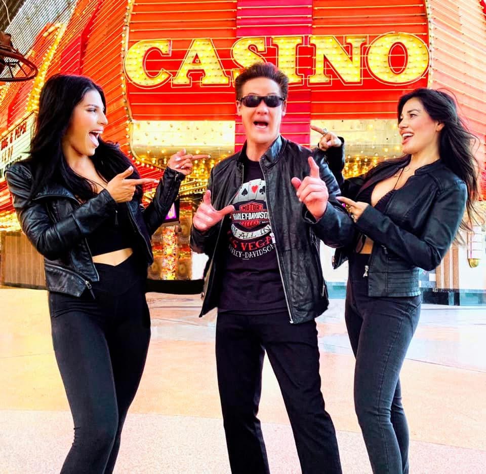 Three people standing in front of a casino, two women and one man, smiling and talking, with bright casino signage behind them.