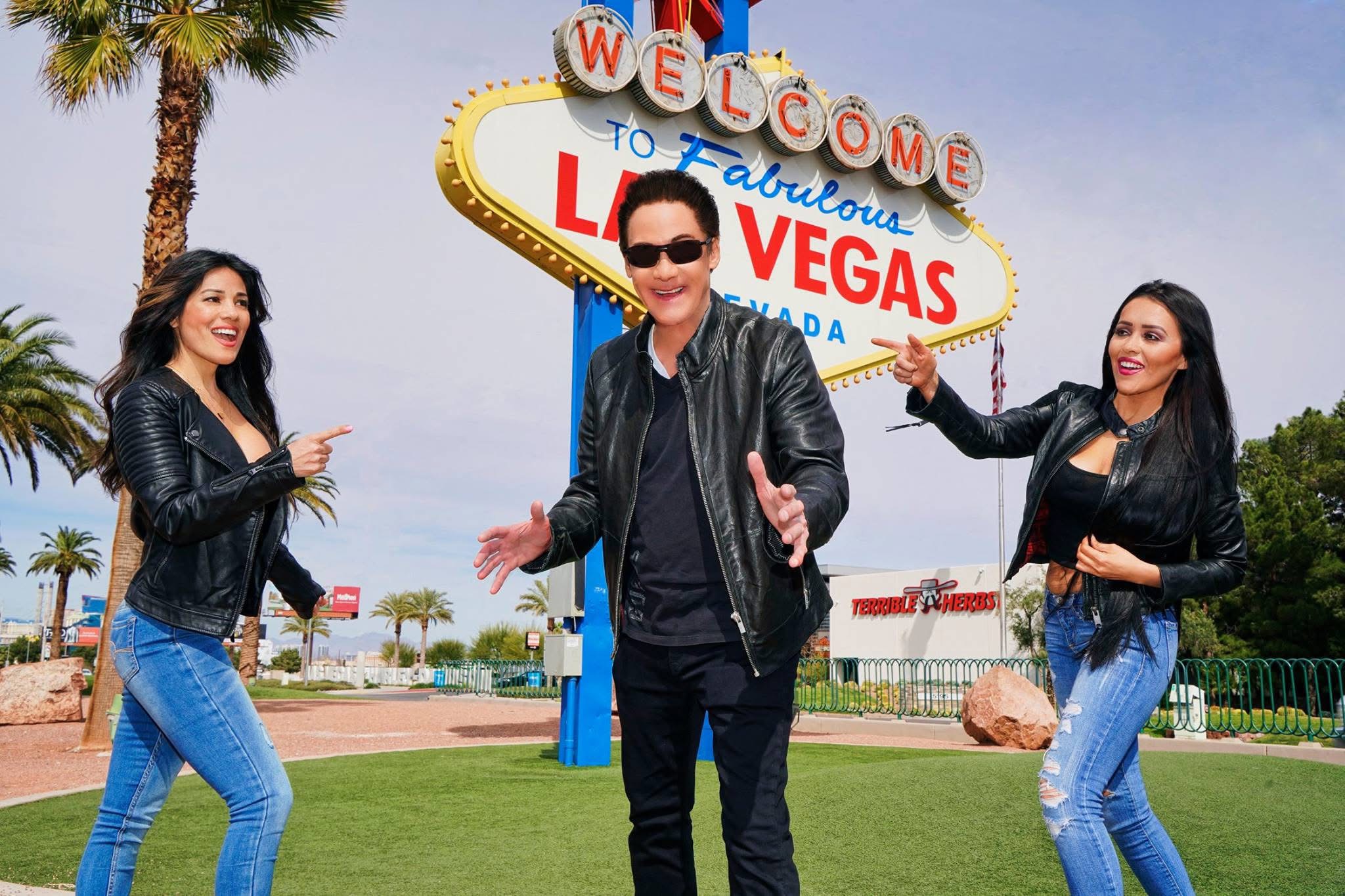 Three people standing outdoors in front of the Las Vegas welcome sign, smiling and pointing at it.