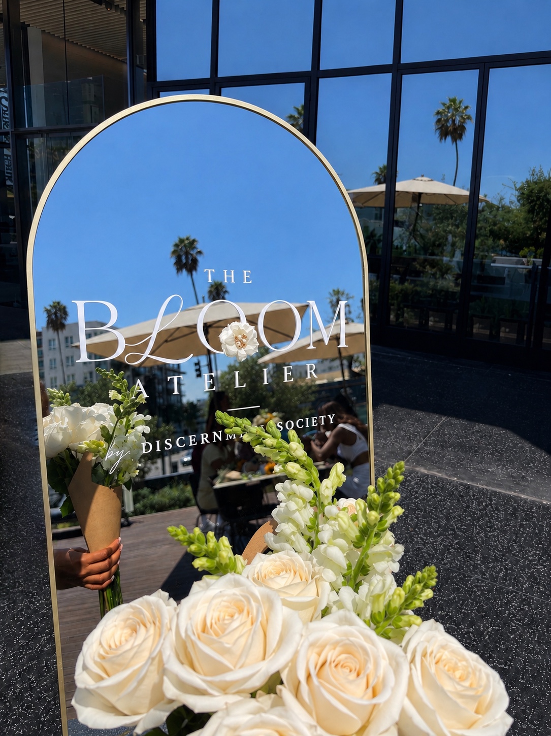 Mirror reflecting outdoor scene with palm trees, blue sky, and a building, with a floral arrangement in front.