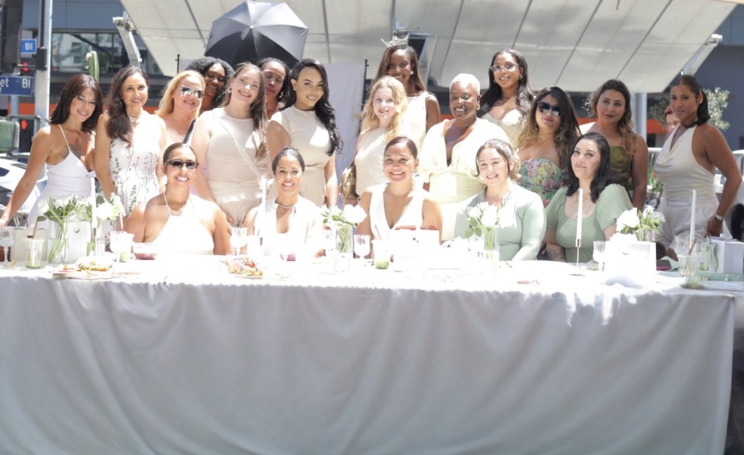 Group of women in white and light-colored dresses sitting and standing behind a long table outdoors, some holding umbrellas.