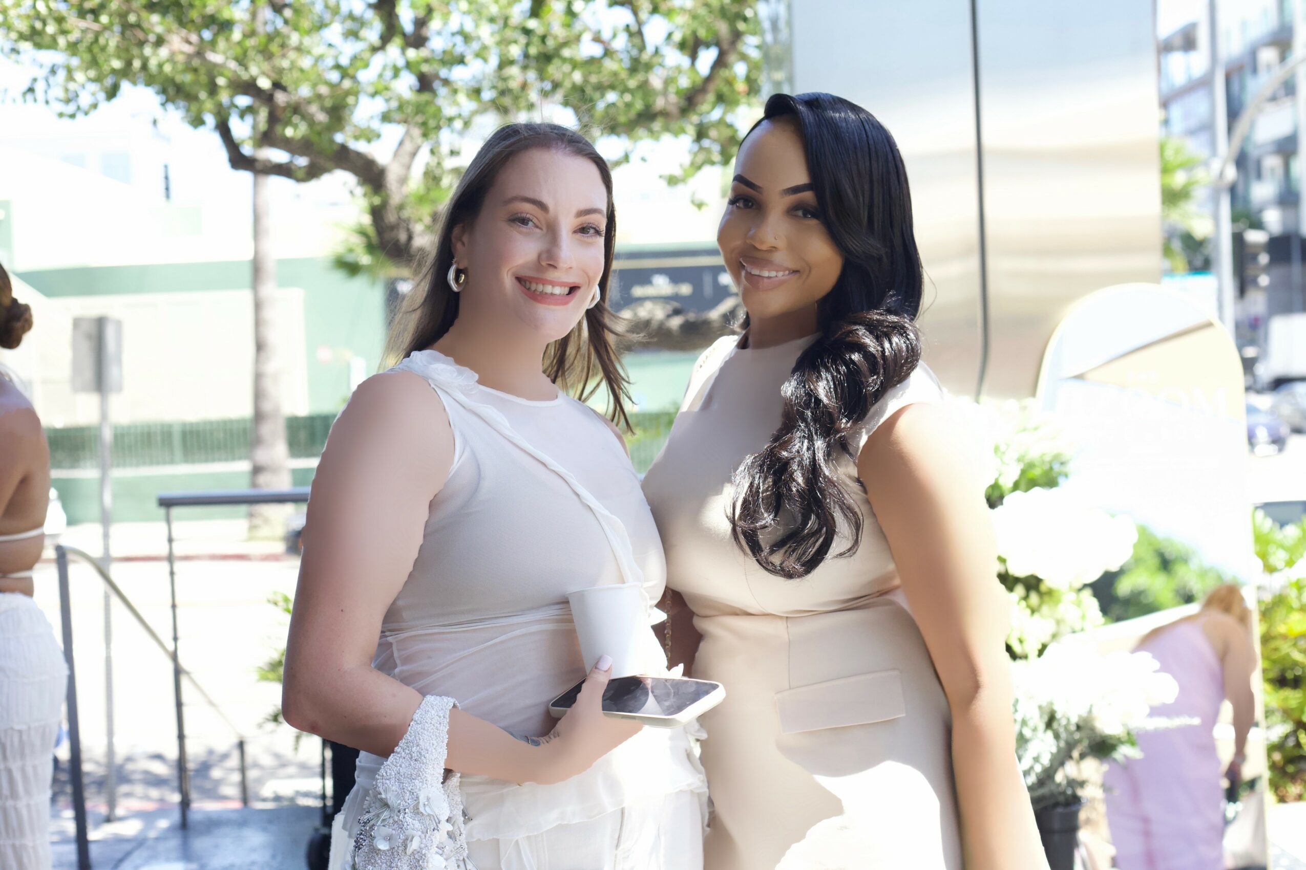 Two women smiling outdoors, one holding a phone, with trees and buildings in the background.