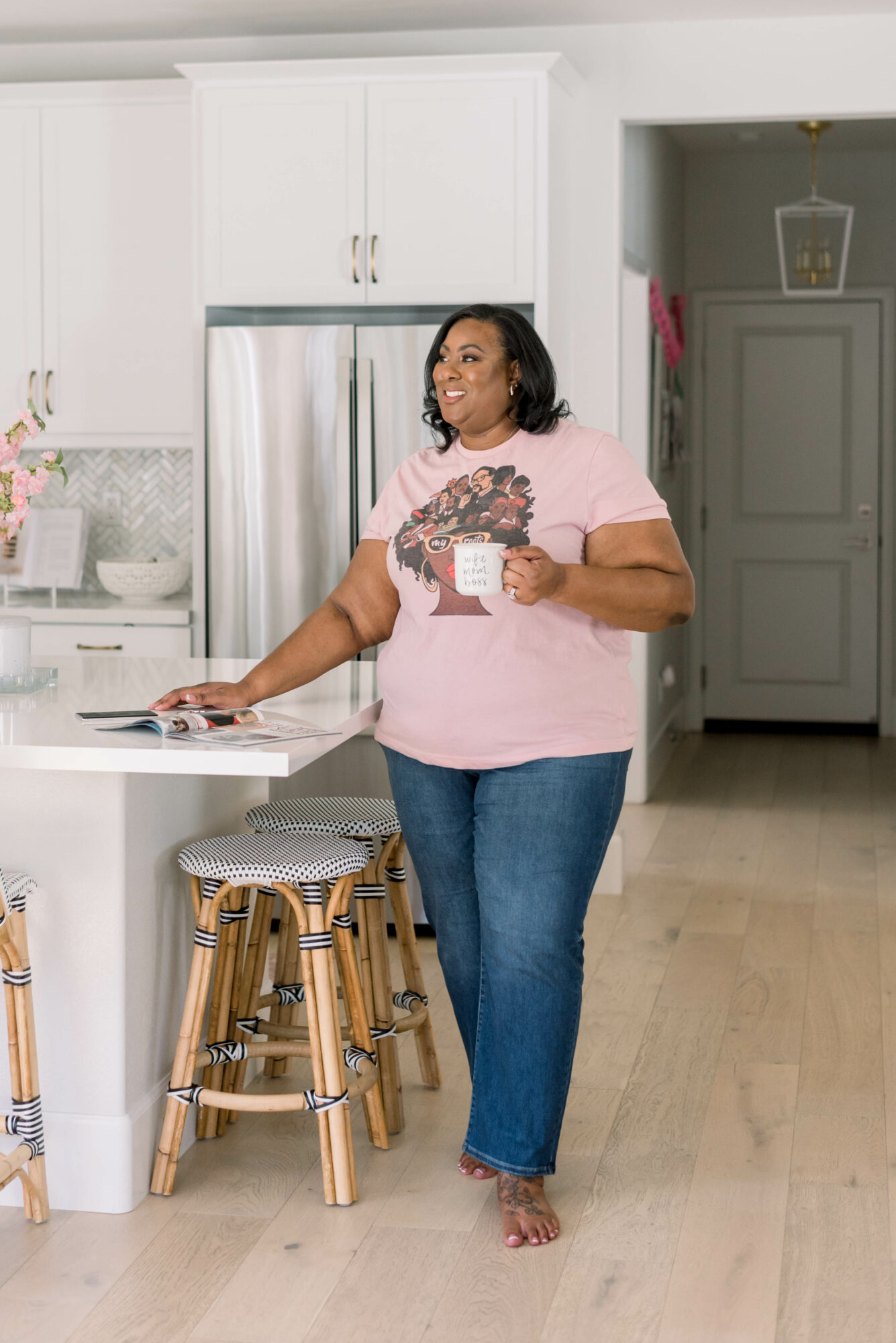 Woman standing in kitchen holding a mug, wearing a pink shirt and jeans, with a refrigerator and cabinets in background.