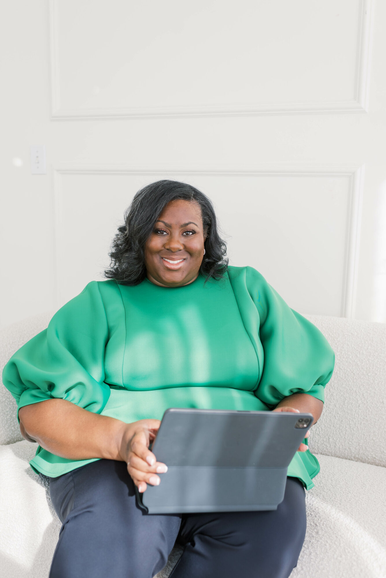 Woman sitting on a white surface holding a tablet, smiling, wearing a green top and dark pants.