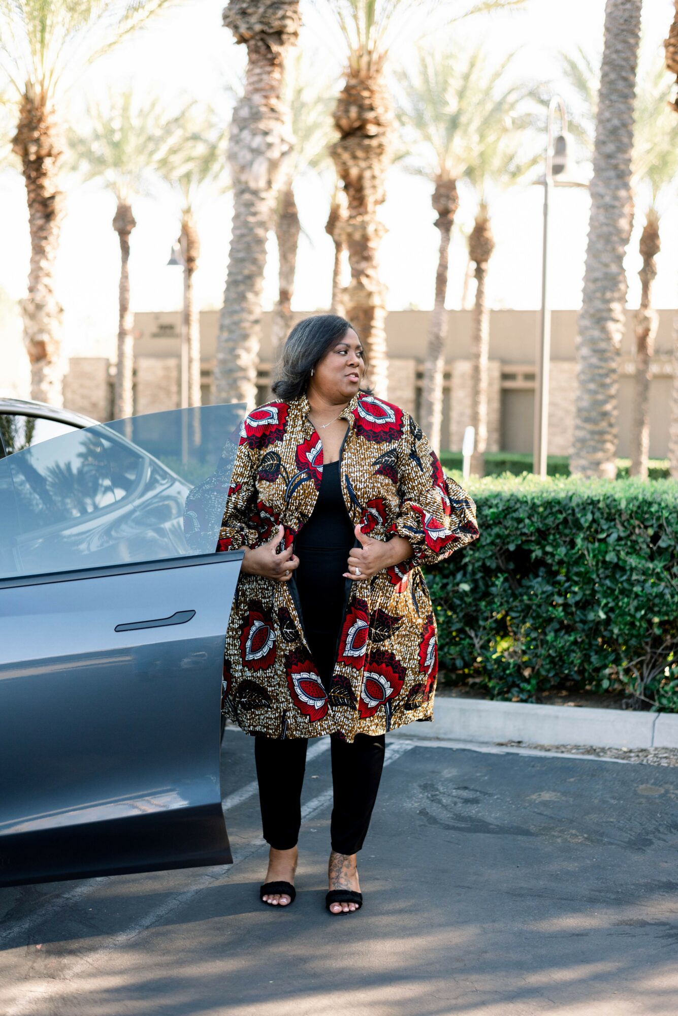 Woman standing outside near a car, wearing a colorful patterned coat and black outfit, with palm trees in background.