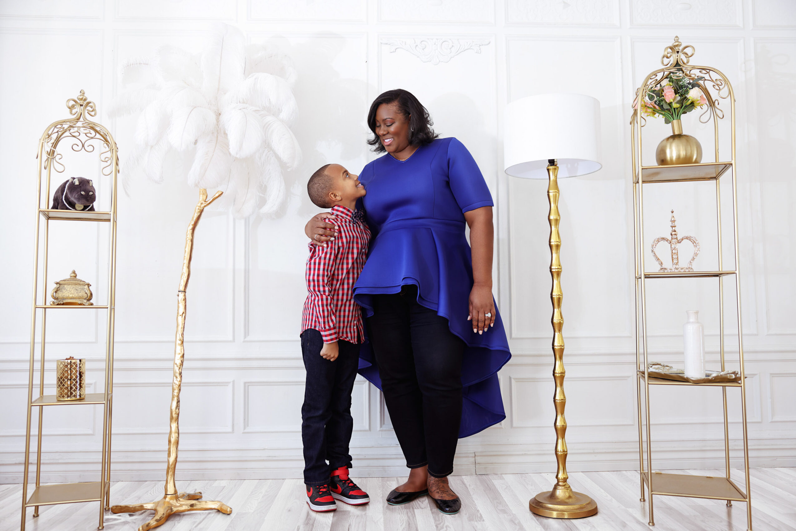 Woman and child standing in a decorated room with white walls, gold shelves, and a tall lamp, smiling at each other.