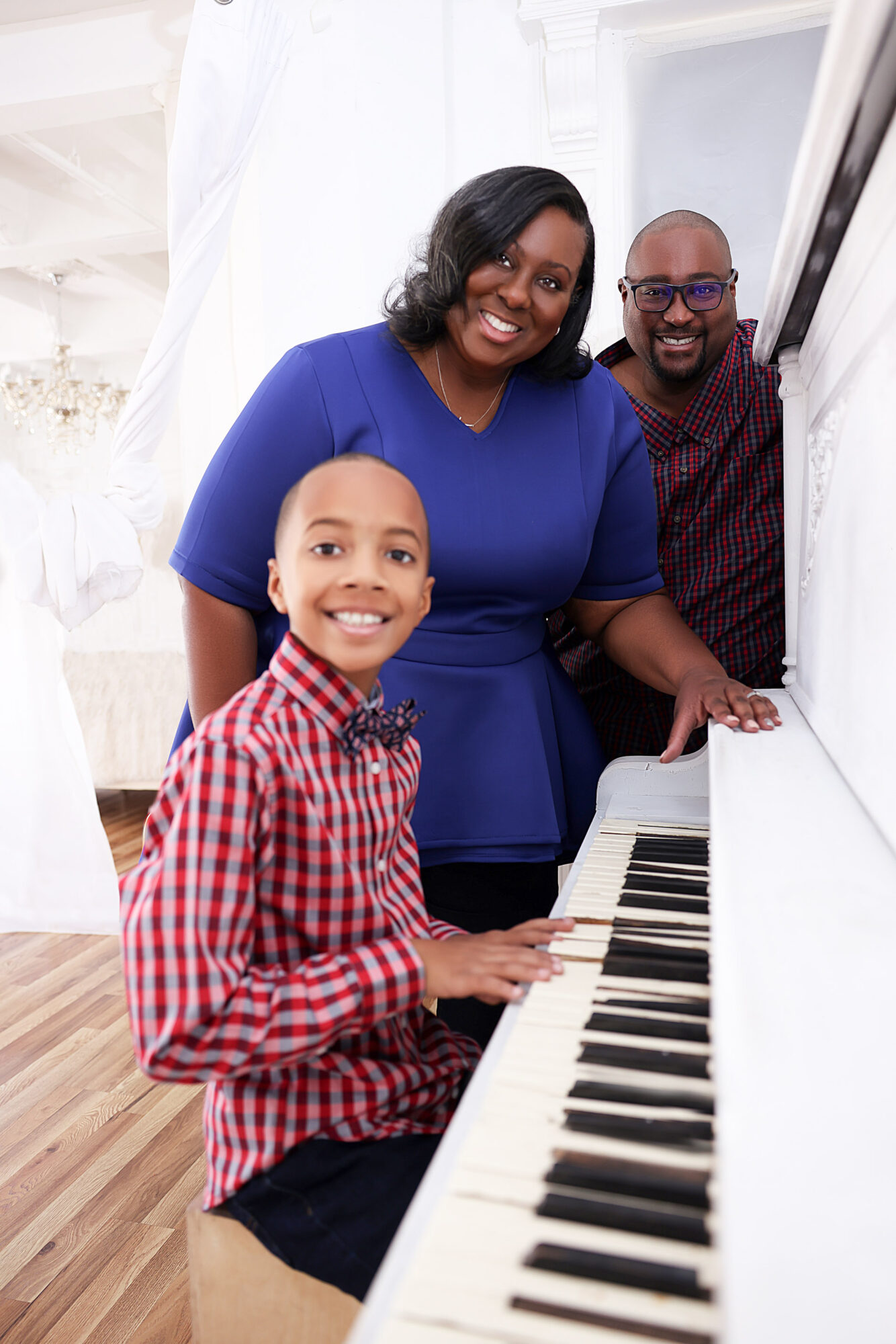 Family gathered around a white piano, smiling, in a bright room with white curtains and wooden floor.