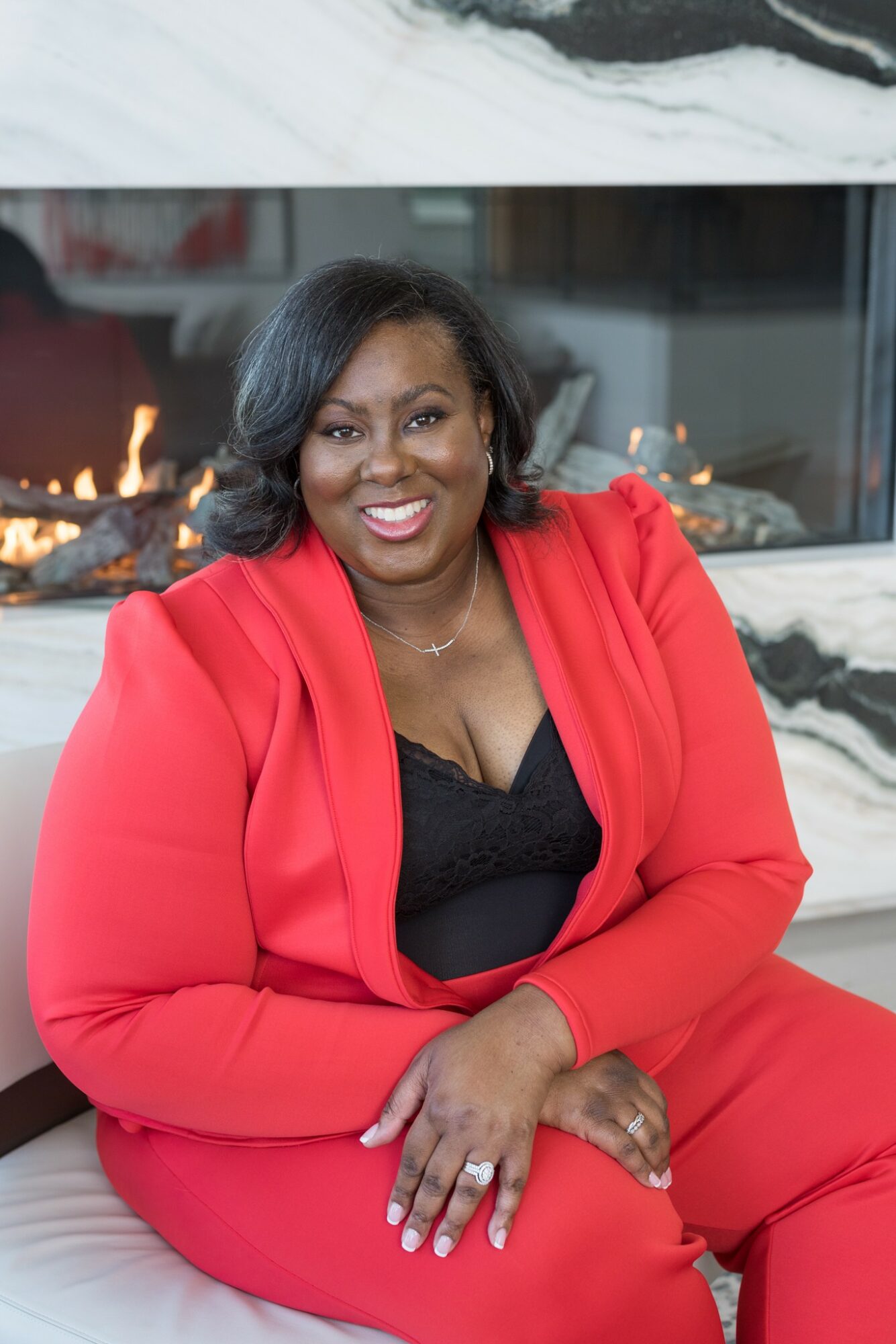 Woman in red suit sitting on a white surface, smiling, with a fireplace in the background.