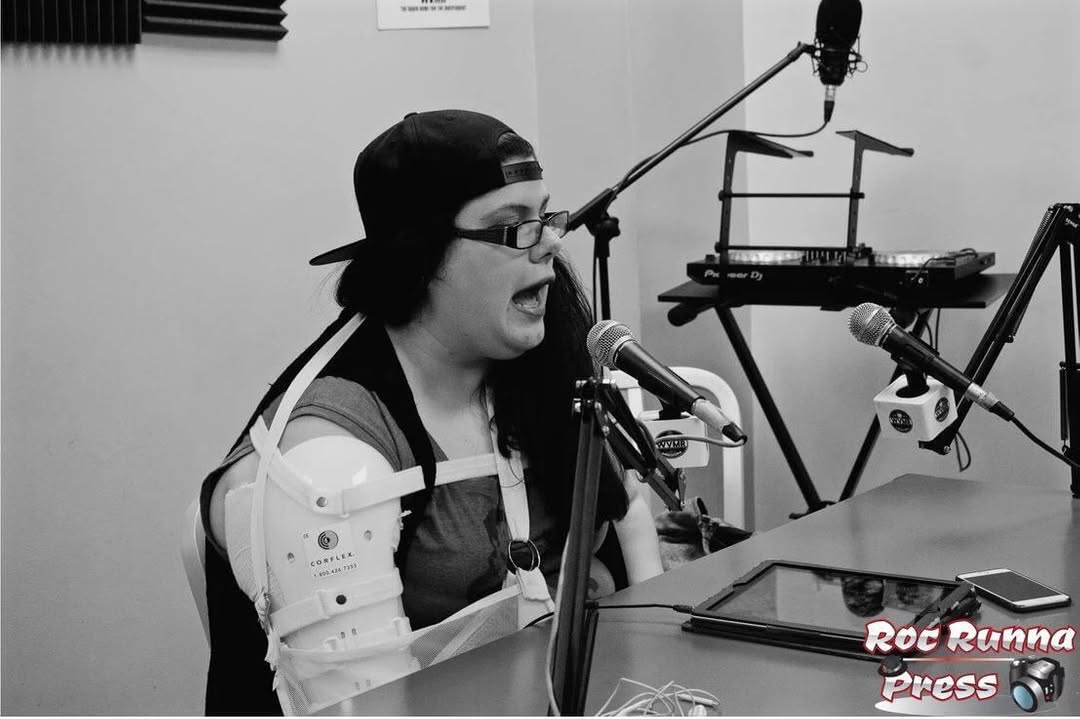 Person singing into a microphone with musical equipment in a recording studio, wearing glasses and a cap.