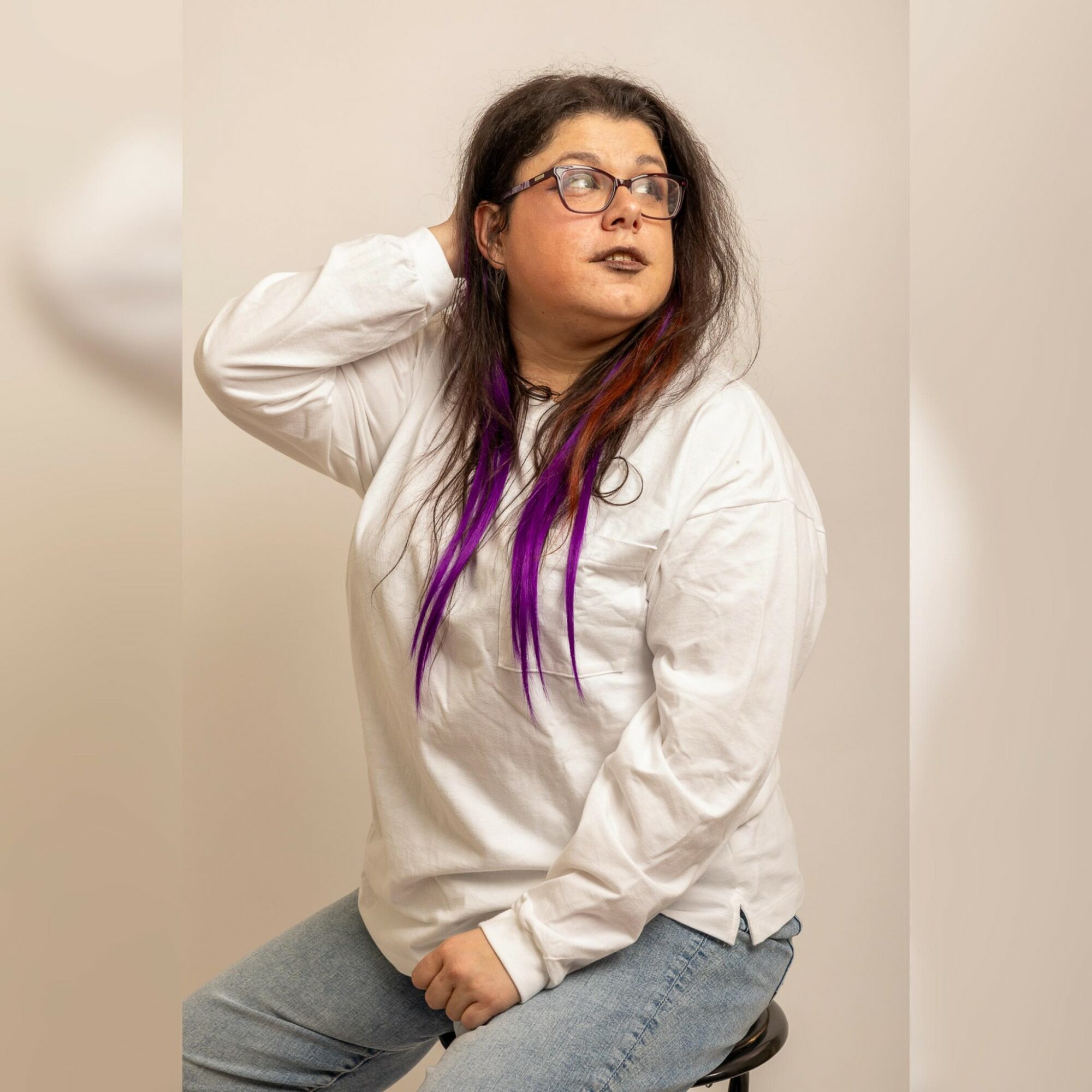 Woman with glasses and long hair with purple streaks, wearing a white shirt, sitting on a stool, looking to the side.