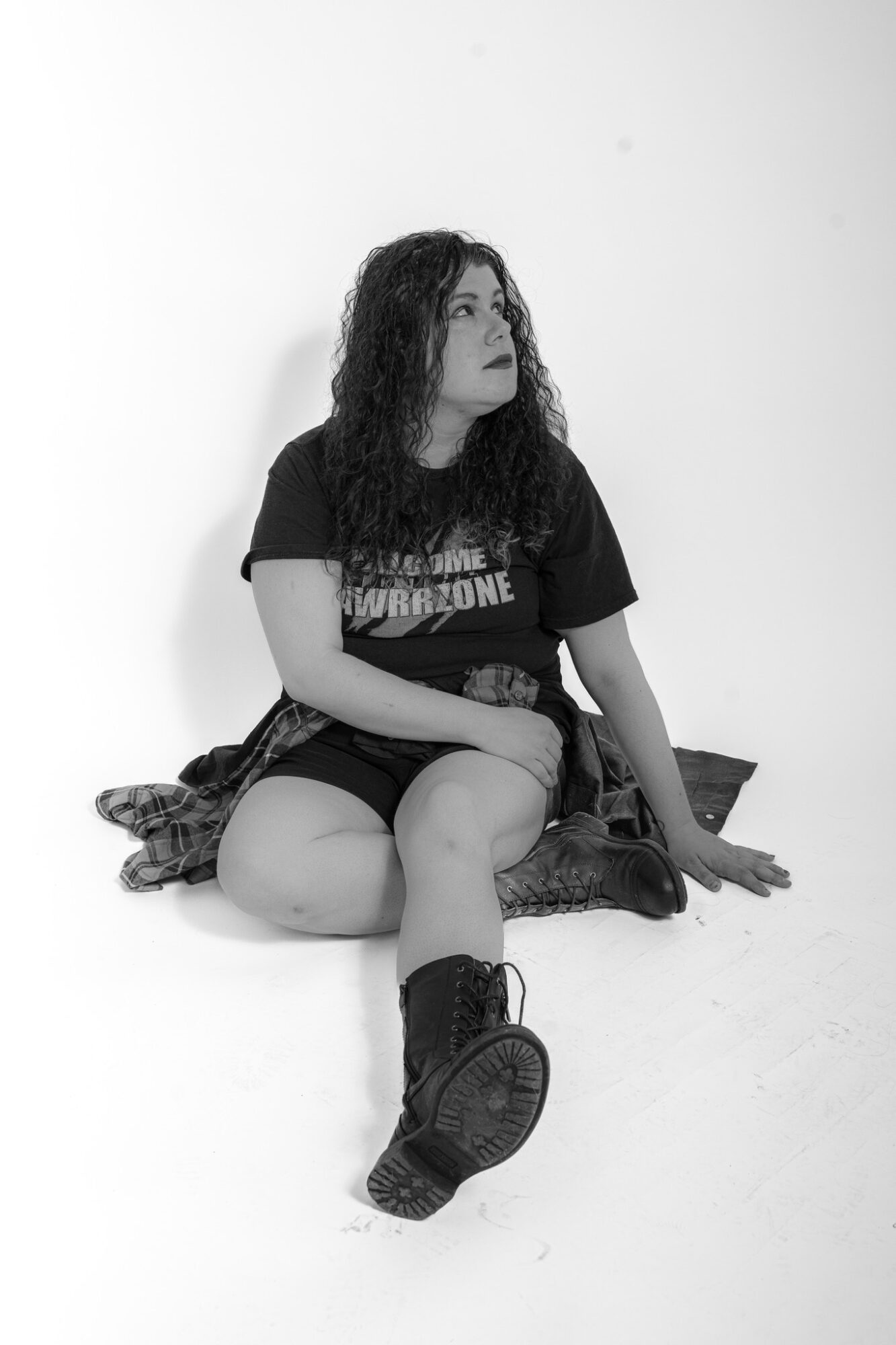 Woman with curly hair sitting on the floor, wearing a T-shirt, shorts, and boots, against a plain background.