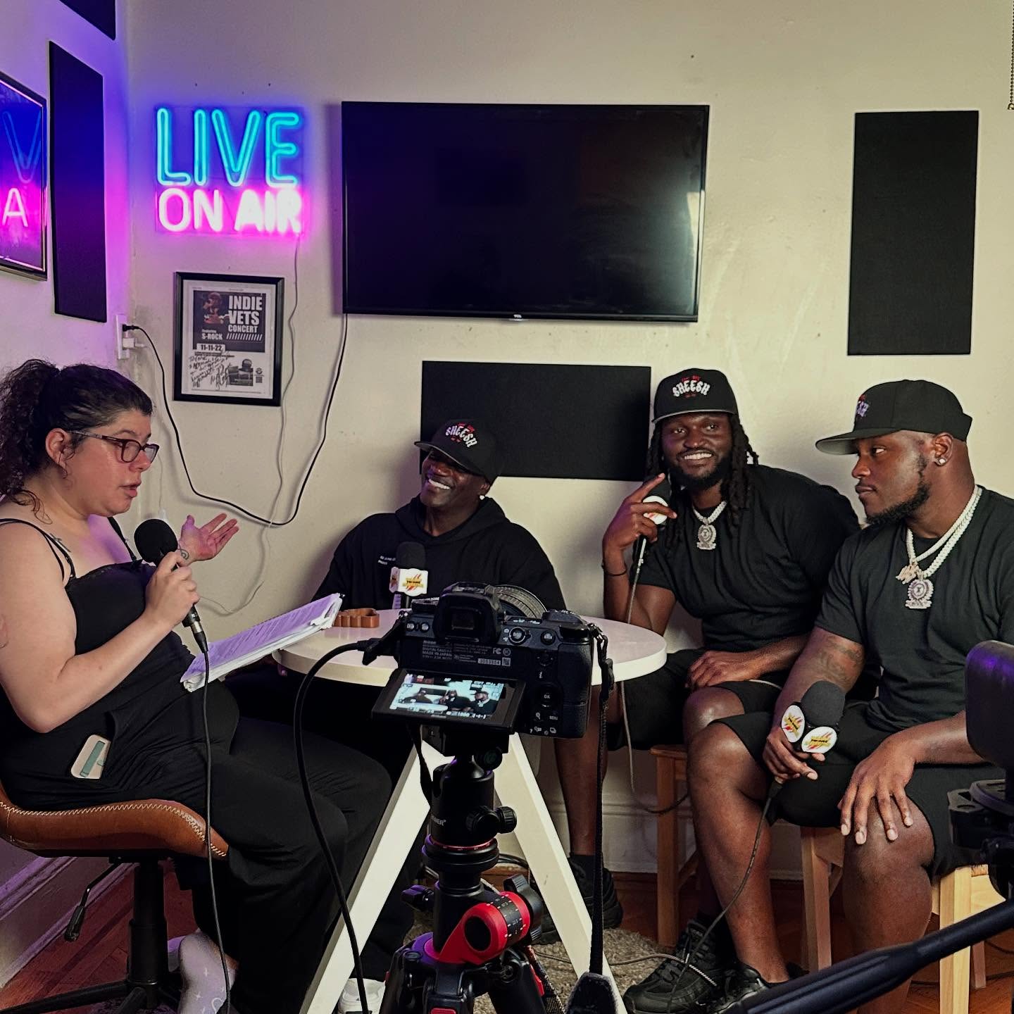 Four people in a radio studio, one woman with a microphone and three men with microphones, a TV screen, and a neon sign that says 'LIVE ON AIR'.