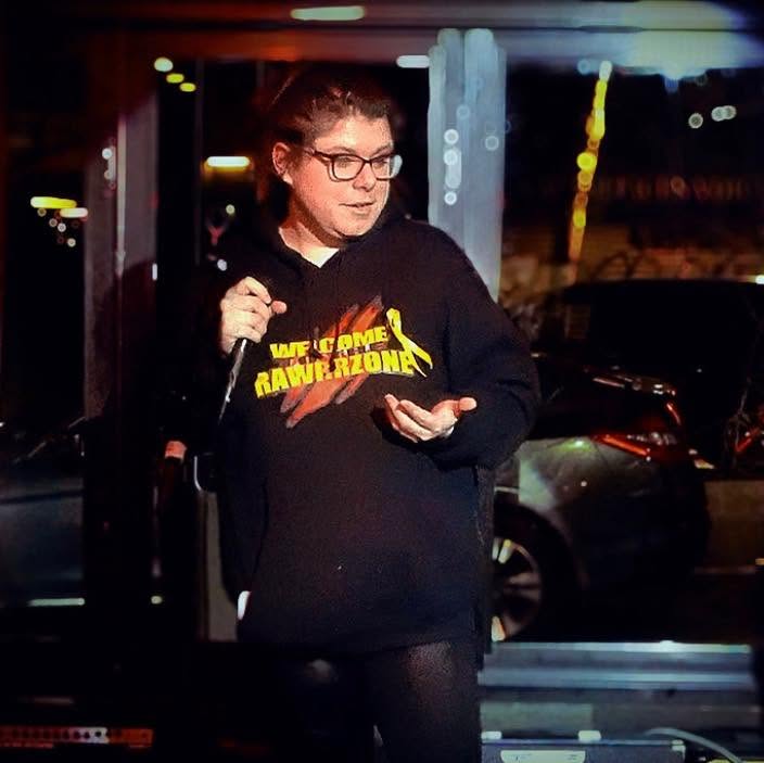 Woman with glasses holding a microphone, wearing a black hoodie with yellow text, standing outdoors at night.