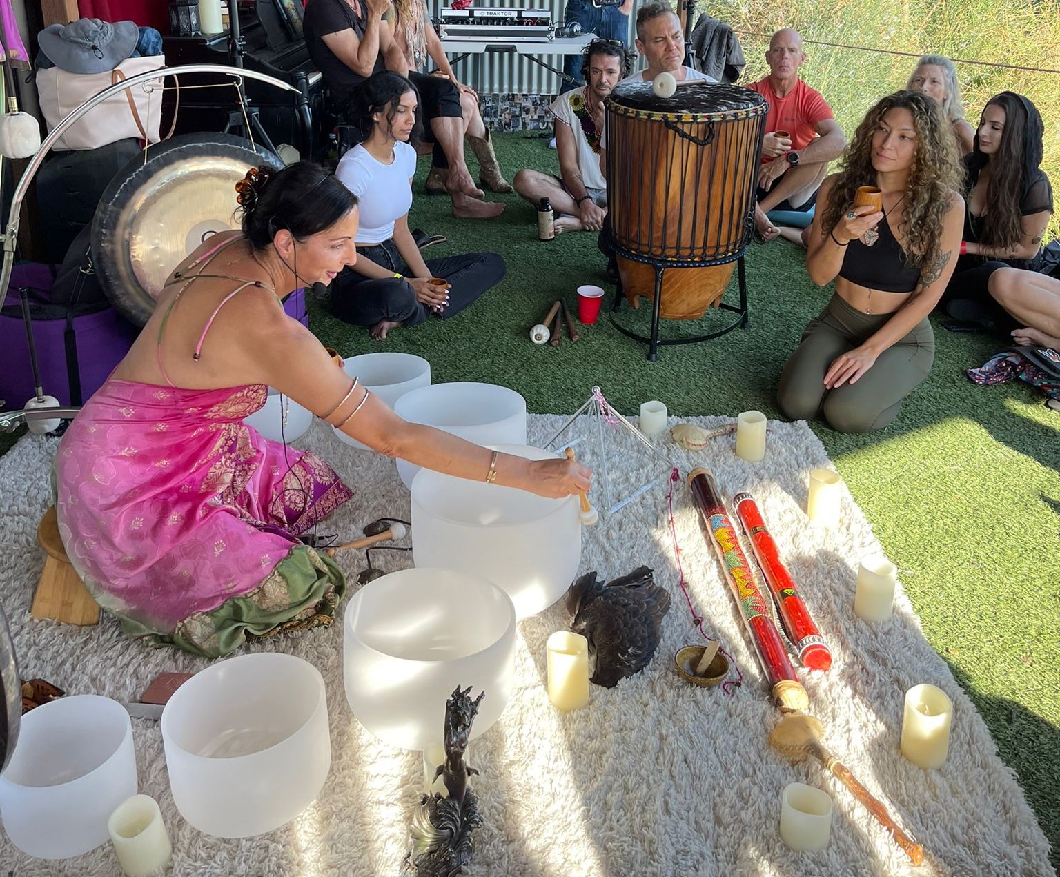 Group of people sitting outdoors, some playing musical instruments, with candles and singing bowls on the ground.