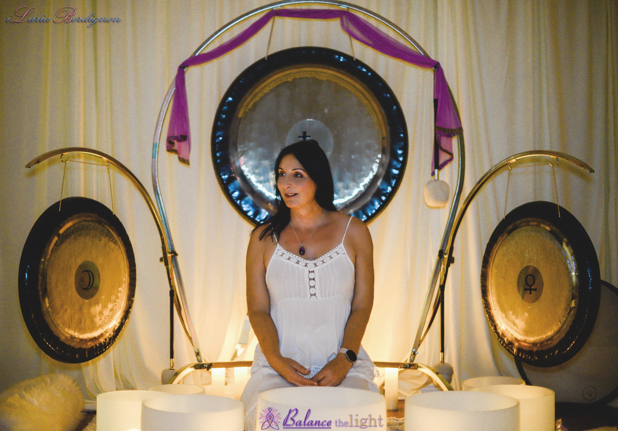 Woman in white dress standing in front of large gong and musical instruments, with curtains in background.