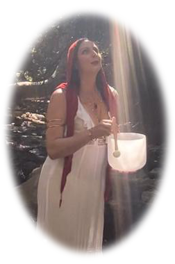 Woman in white dress holding a singing bowl outdoors with sunlight shining on her, trees in background.