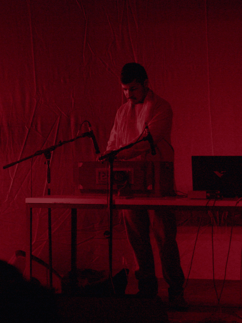 Person standing at a table with electronic equipment, red lighting, and microphones, performing or presenting on stage.