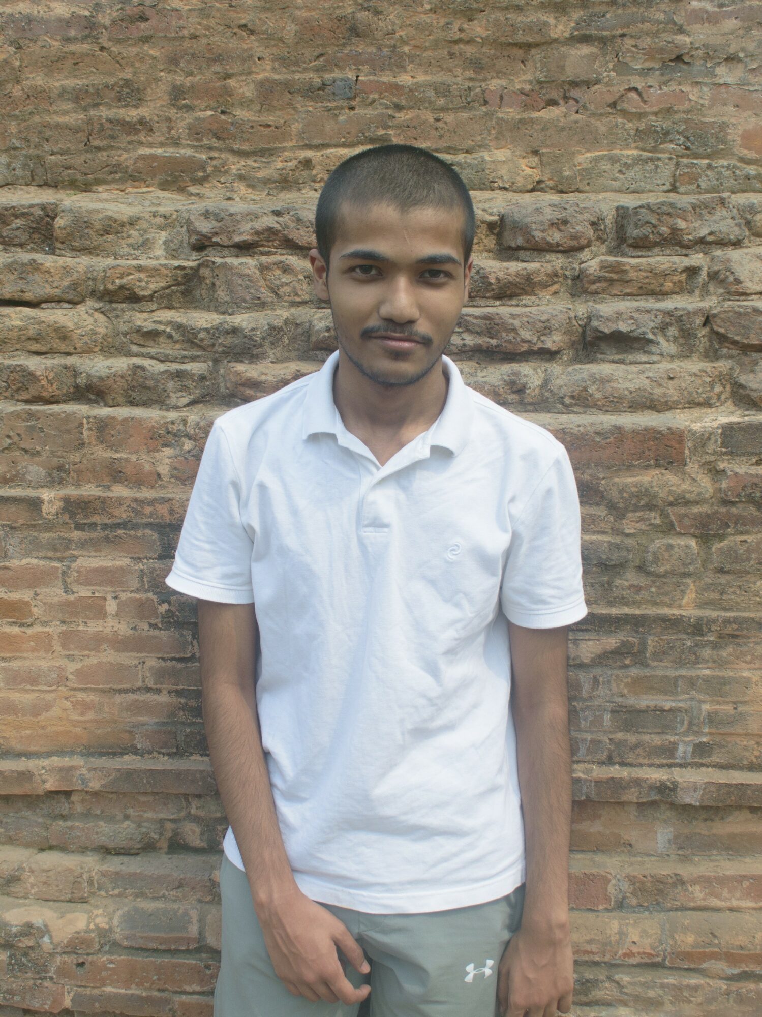 Young person with short hair wearing a white shirt standing against a brick wall.
