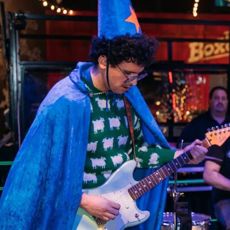Person wearing a blue cape and a party hat playing an electric guitar, with a band in the background.