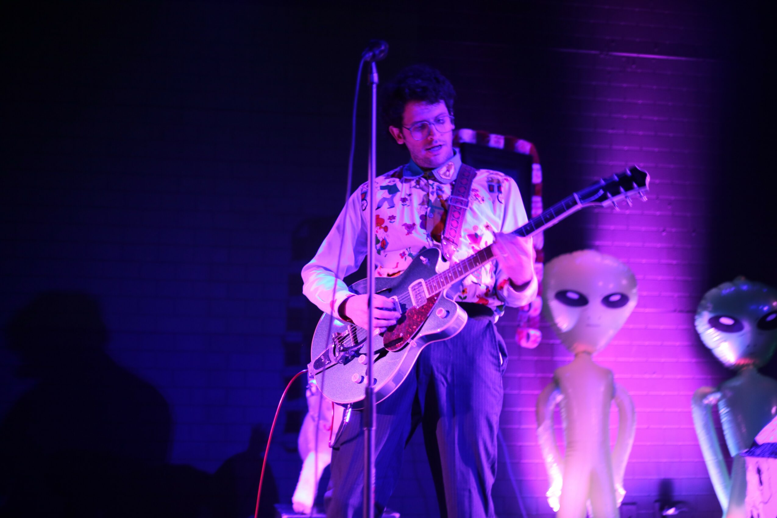 Person playing electric guitar on stage with alien figures in background, purple lighting, and microphone stand.