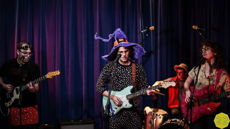 Group of four musicians performing on stage with colorful costumes and instruments, dark curtain background.