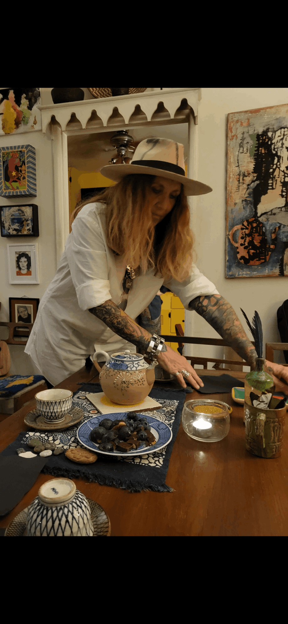 Person with long hair and tattoos wearing a wide-brimmed hat and white shirt, arranging items on a table in an art-filled room.