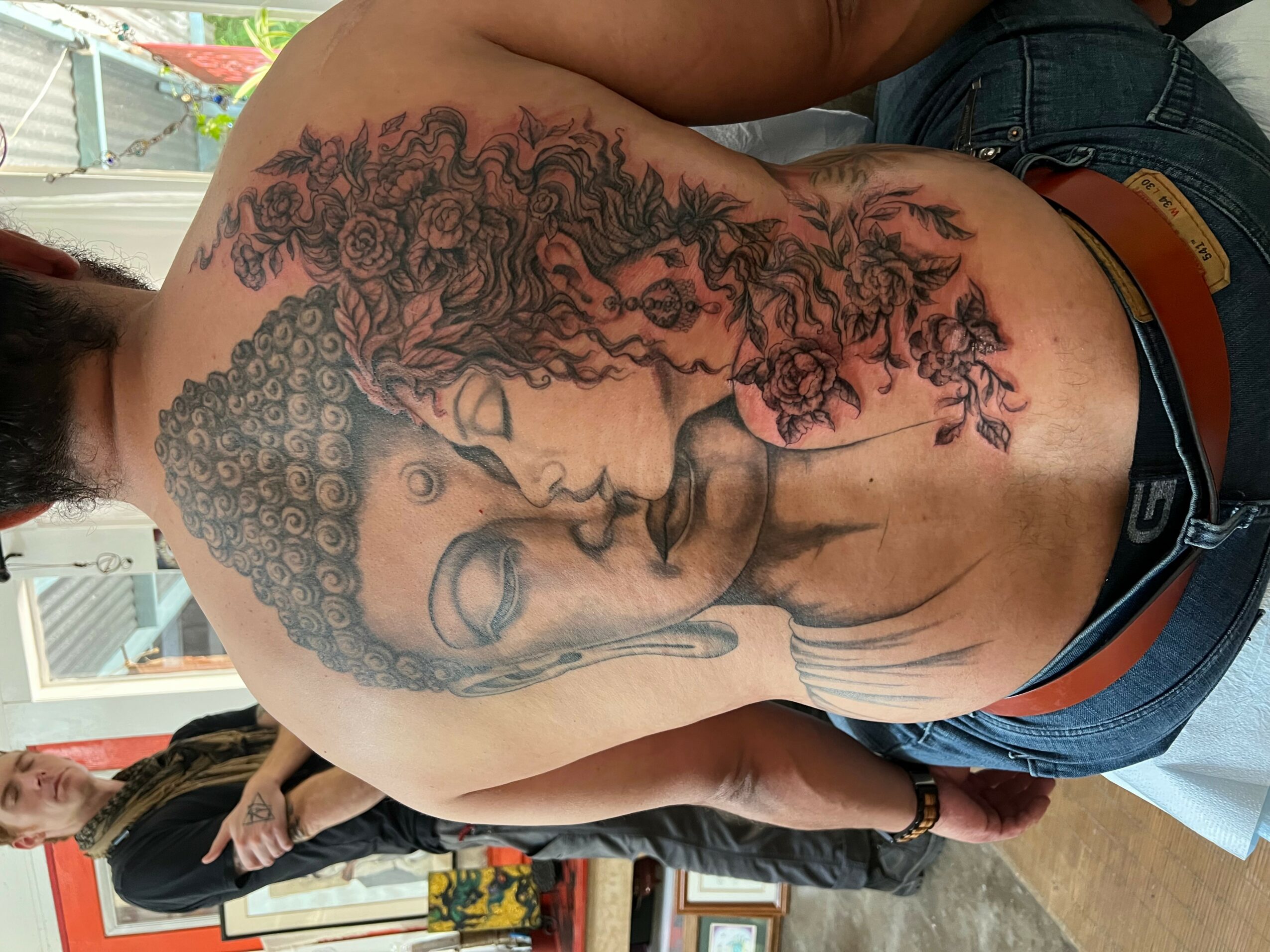 Back view of a person with a detailed tattoo of a woman's face and curly hair on their upper back and shoulder.