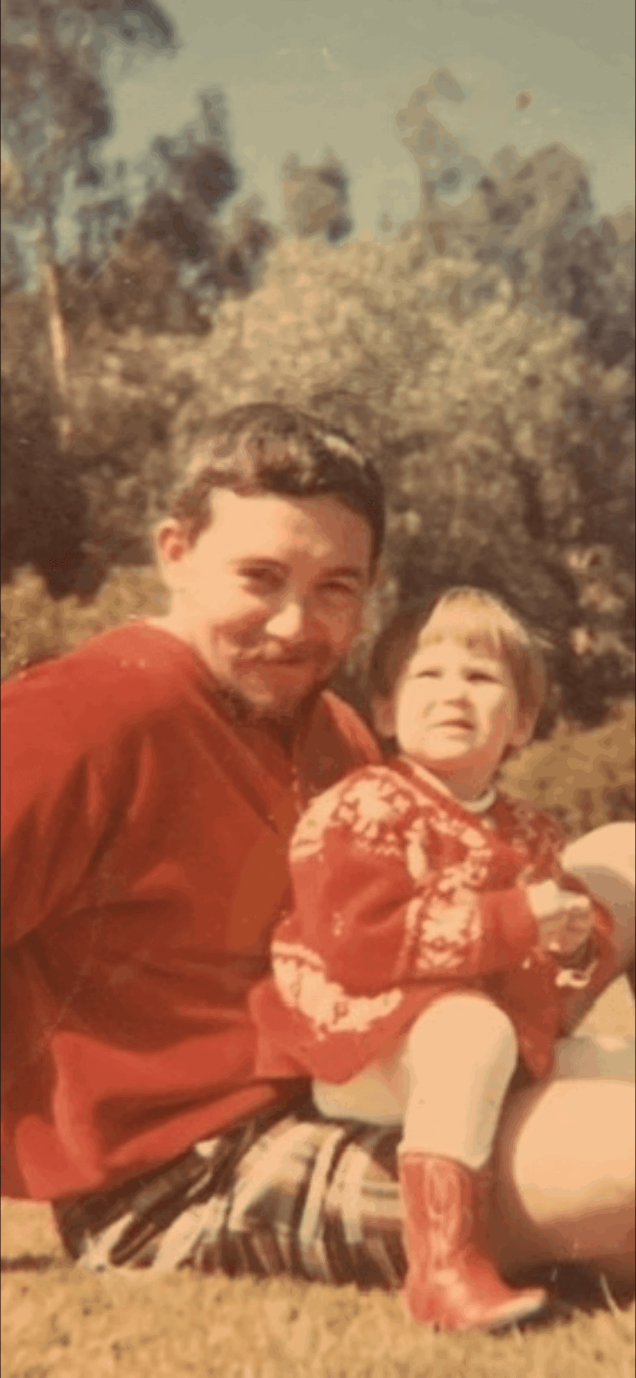 Man holding a young girl outdoors with trees in background, both smiling, girl wearing a red sweater and boots.