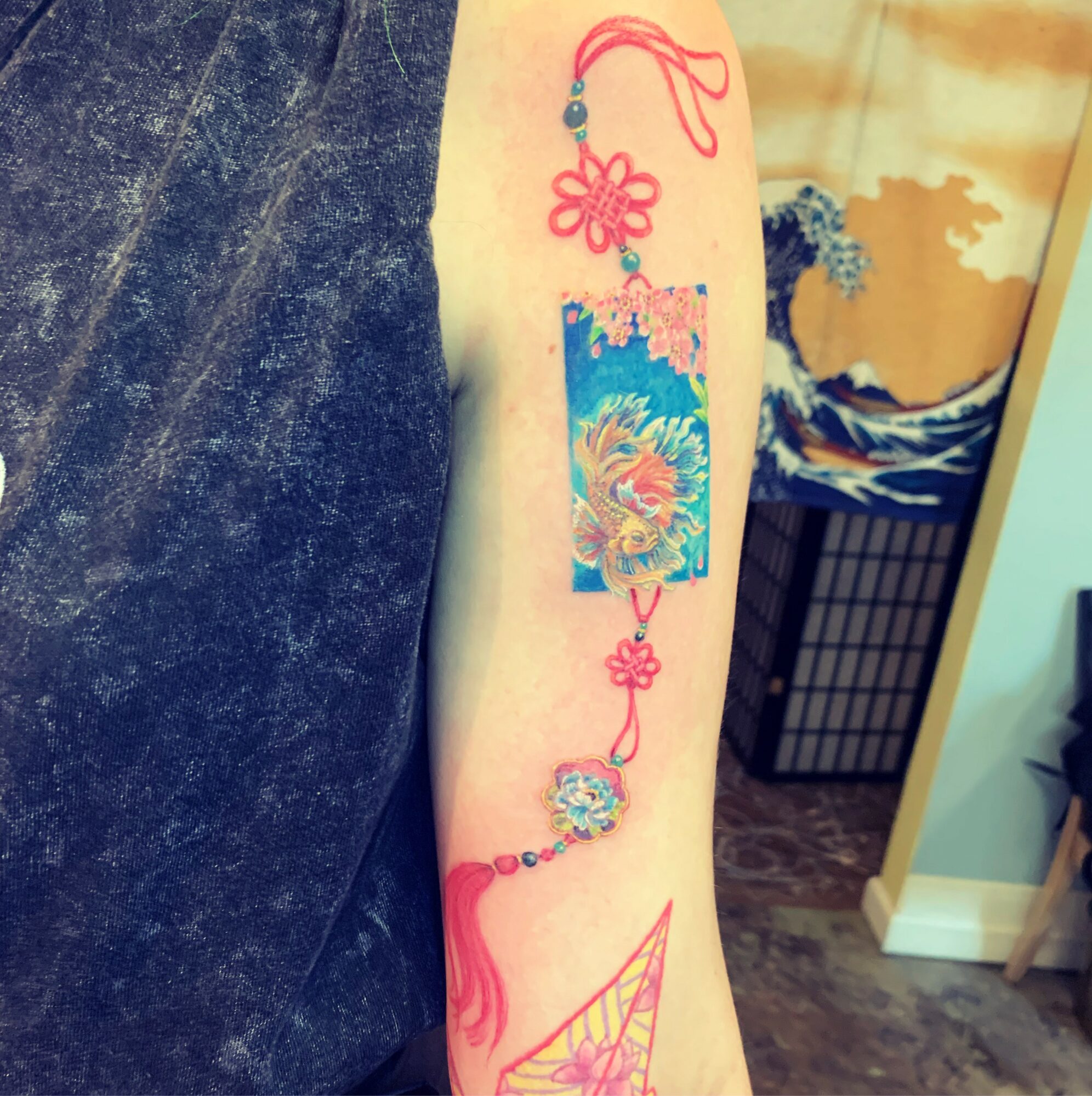 Person's arm with colorful tattoo of a floral design, a rectangular pattern, and decorative elements, against a background of indoor setting.