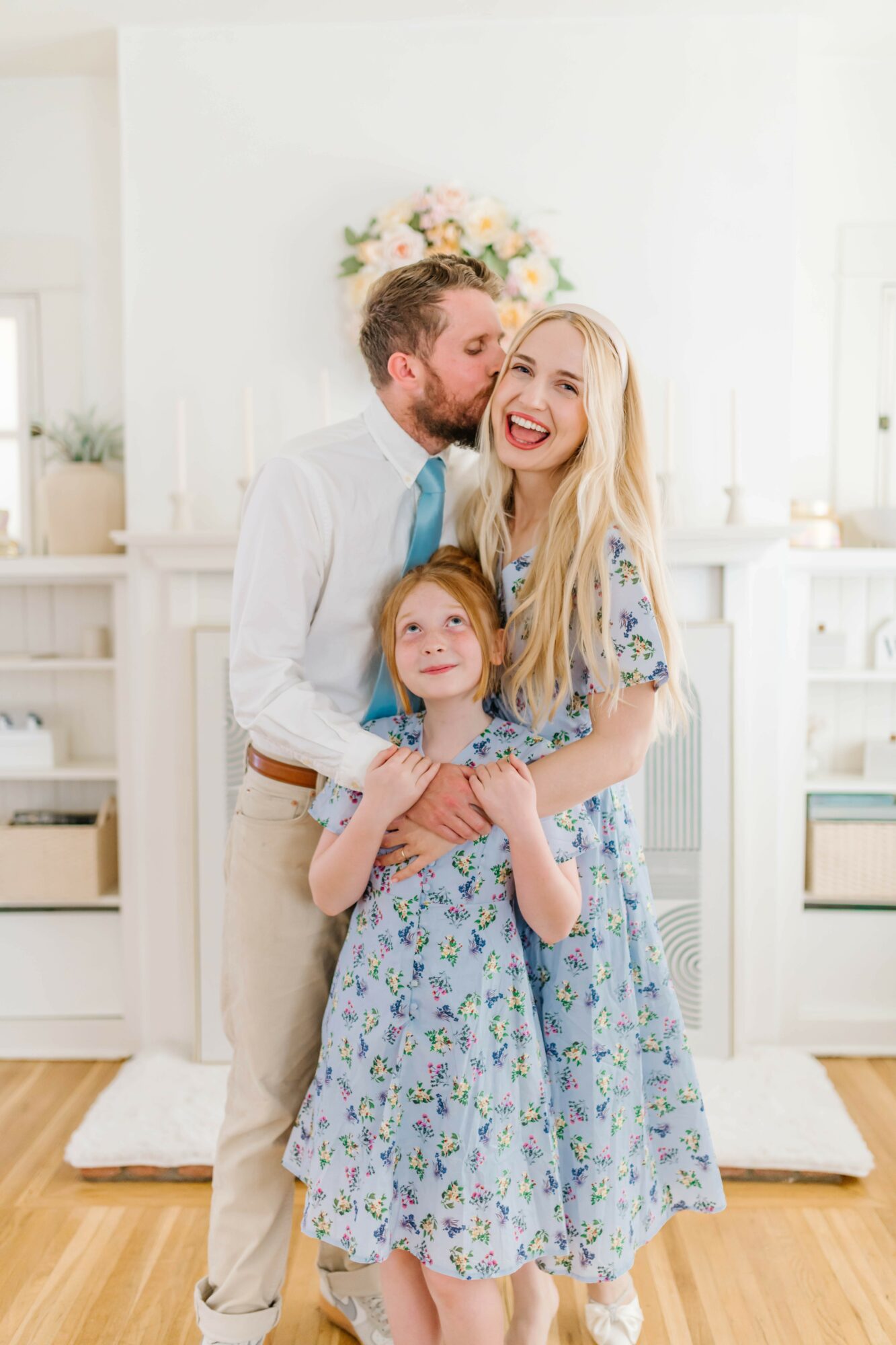 Family of three smiling and hugging in a bright room with white walls and wooden floor, flowers in background.
