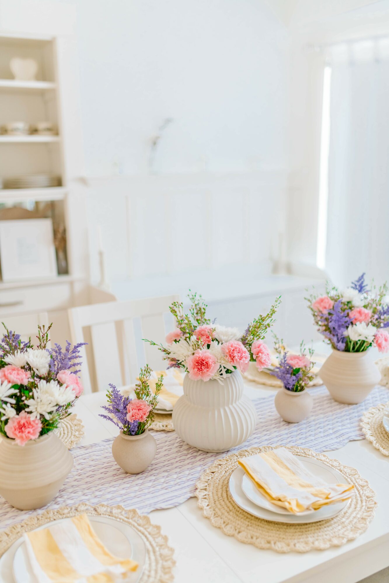 Dining table with white vases holding pink, purple, and green flowers, set with plates and napkins, in a bright room.