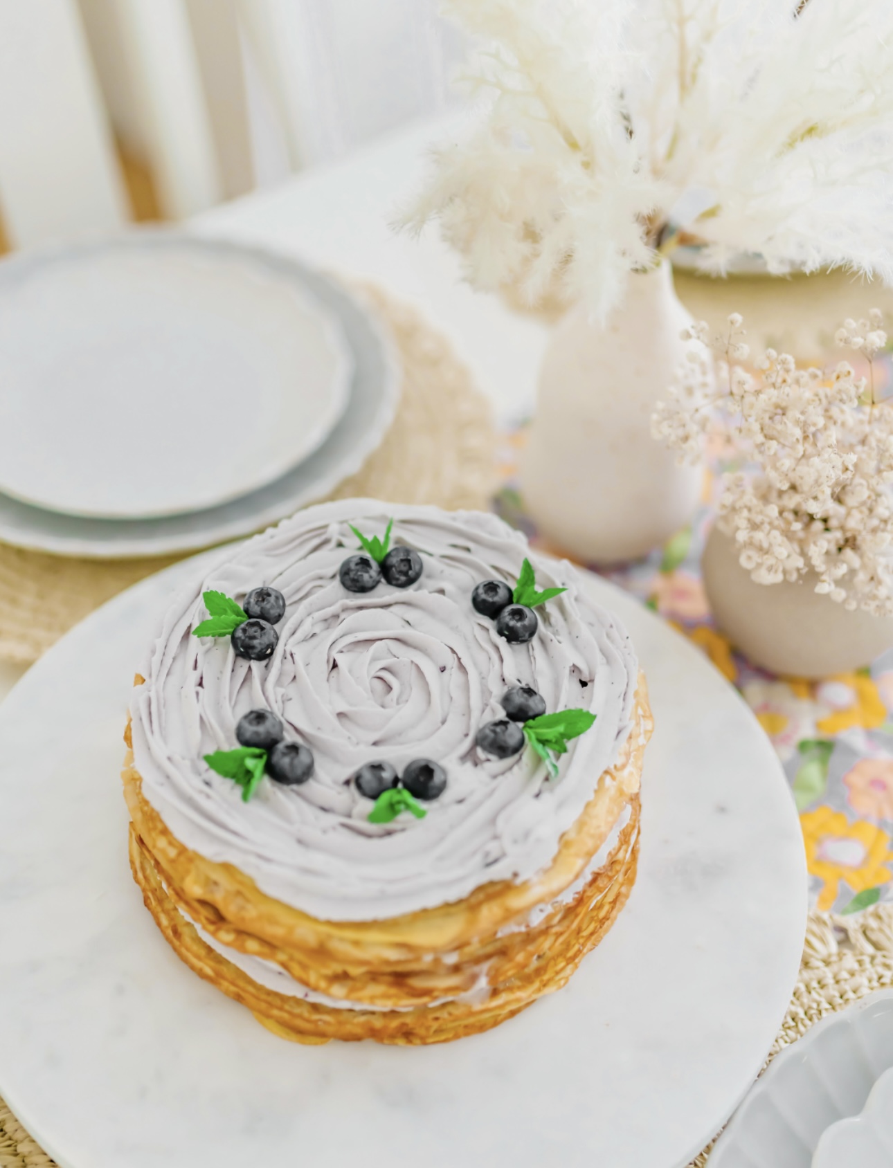 Layered cake with white frosting and blueberries, decorated with green leaves, on a white plate, with vases and flowers in background.