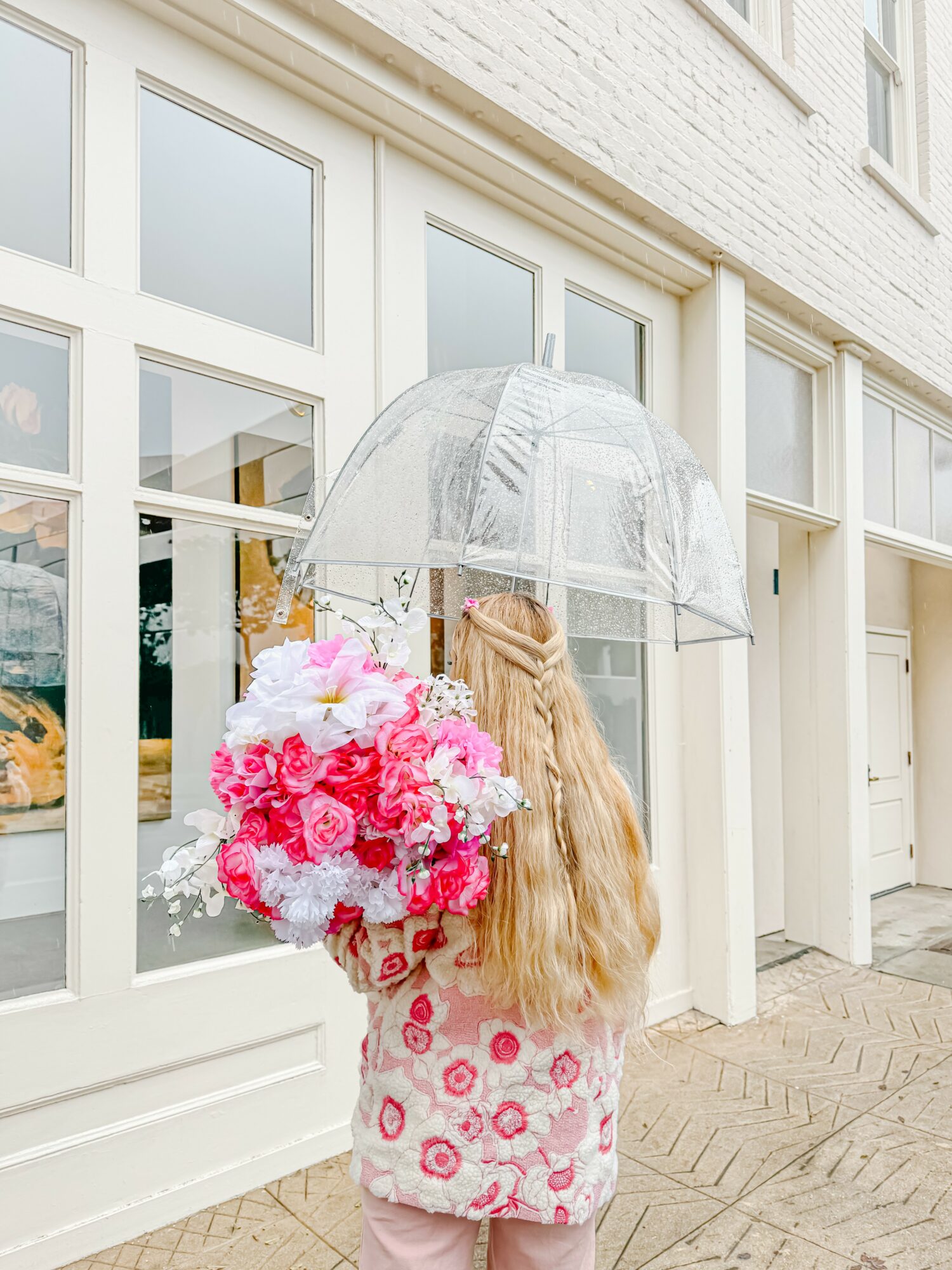 Person with long blonde hair holding a large pink and white flower bouquet under a transparent umbrella outside a building with large windows.
