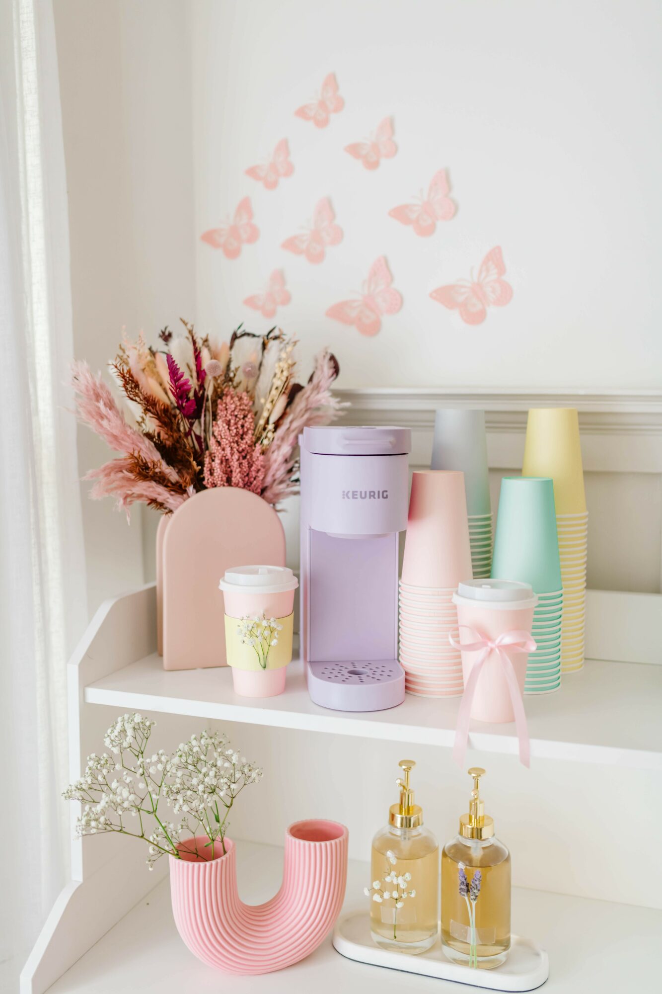 Decorative shelf with pastel cups, bottles, a pink vase with flowers, and pink butterfly wall decals, against a white wall.