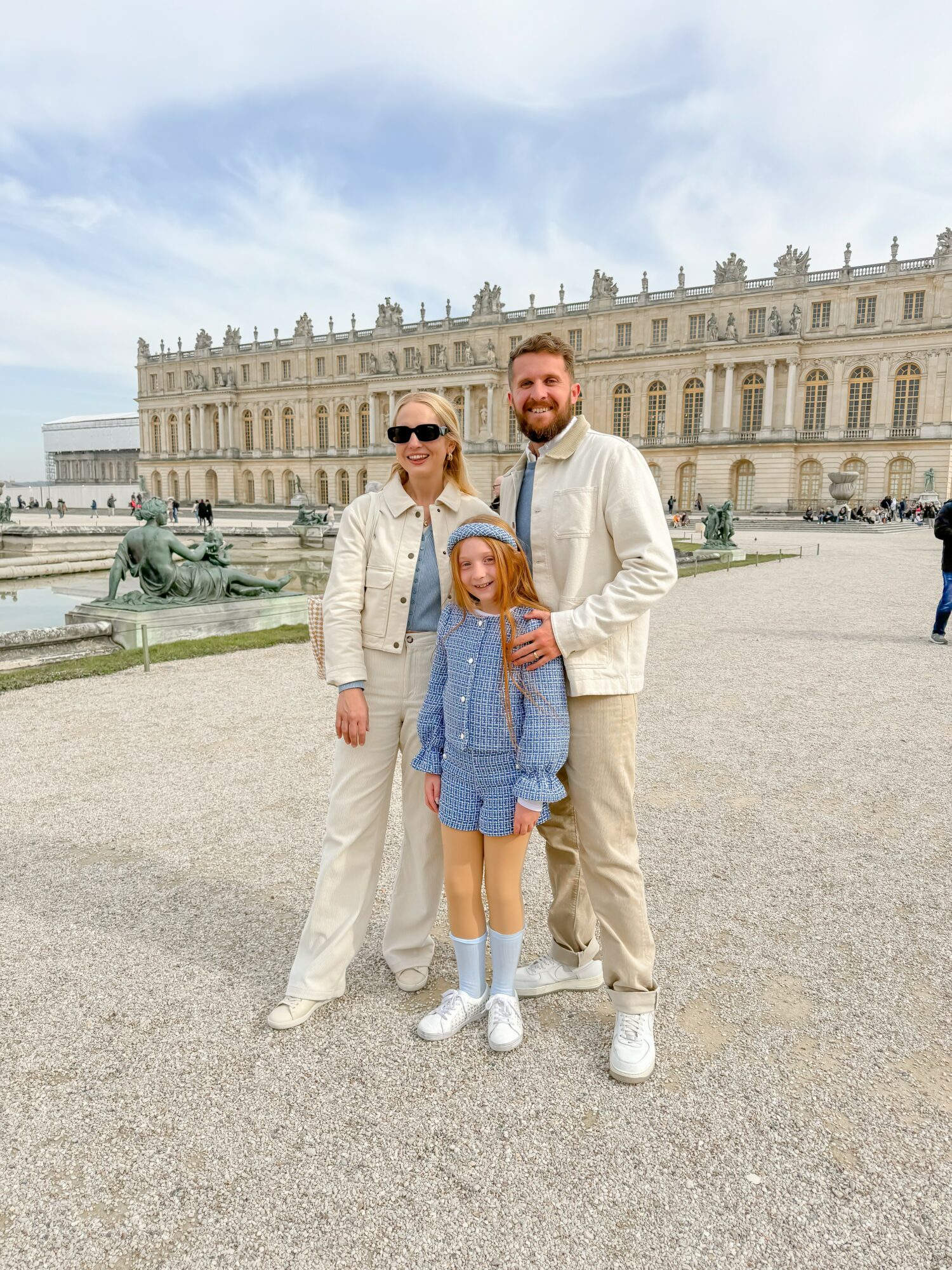 Three people, two adults and a child, stand in front of a historic building with fountains, smiling outdoors.