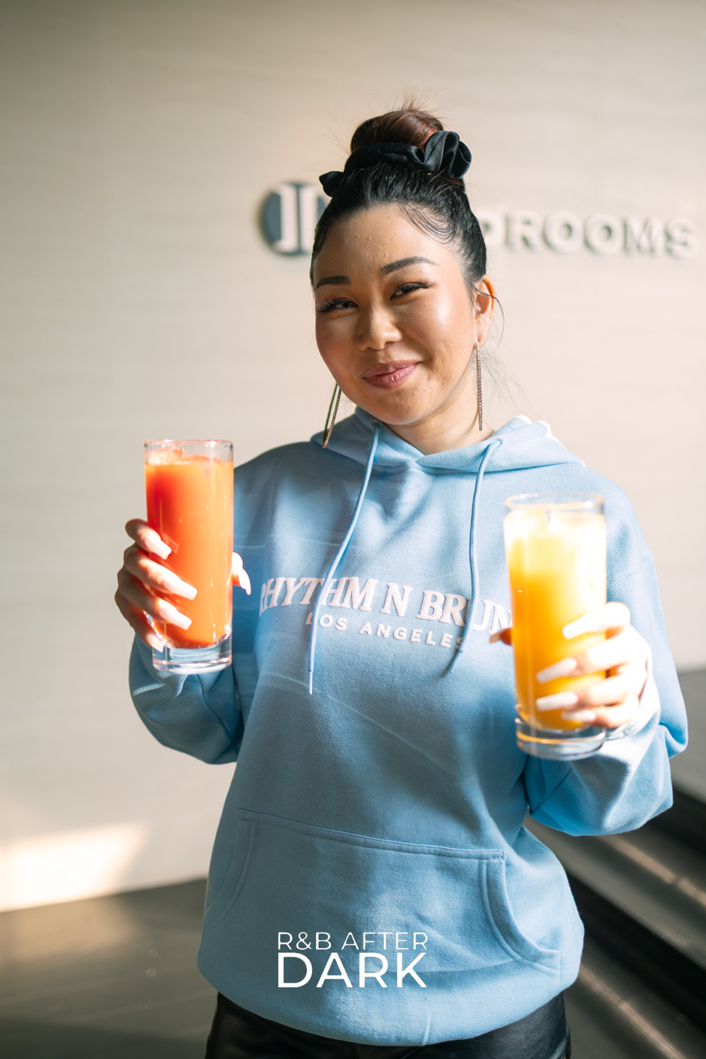 Young woman with dark hair in a bun, smiling, holding two glasses of orange and yellow drinks, wearing a light blue hoodie.